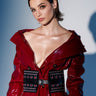 A woman with short, slicked-back hair wears a glossy red trench coat styled with the LOVER GROMMET PU BELT. She poses confidently against a light background with dramatic shadows.