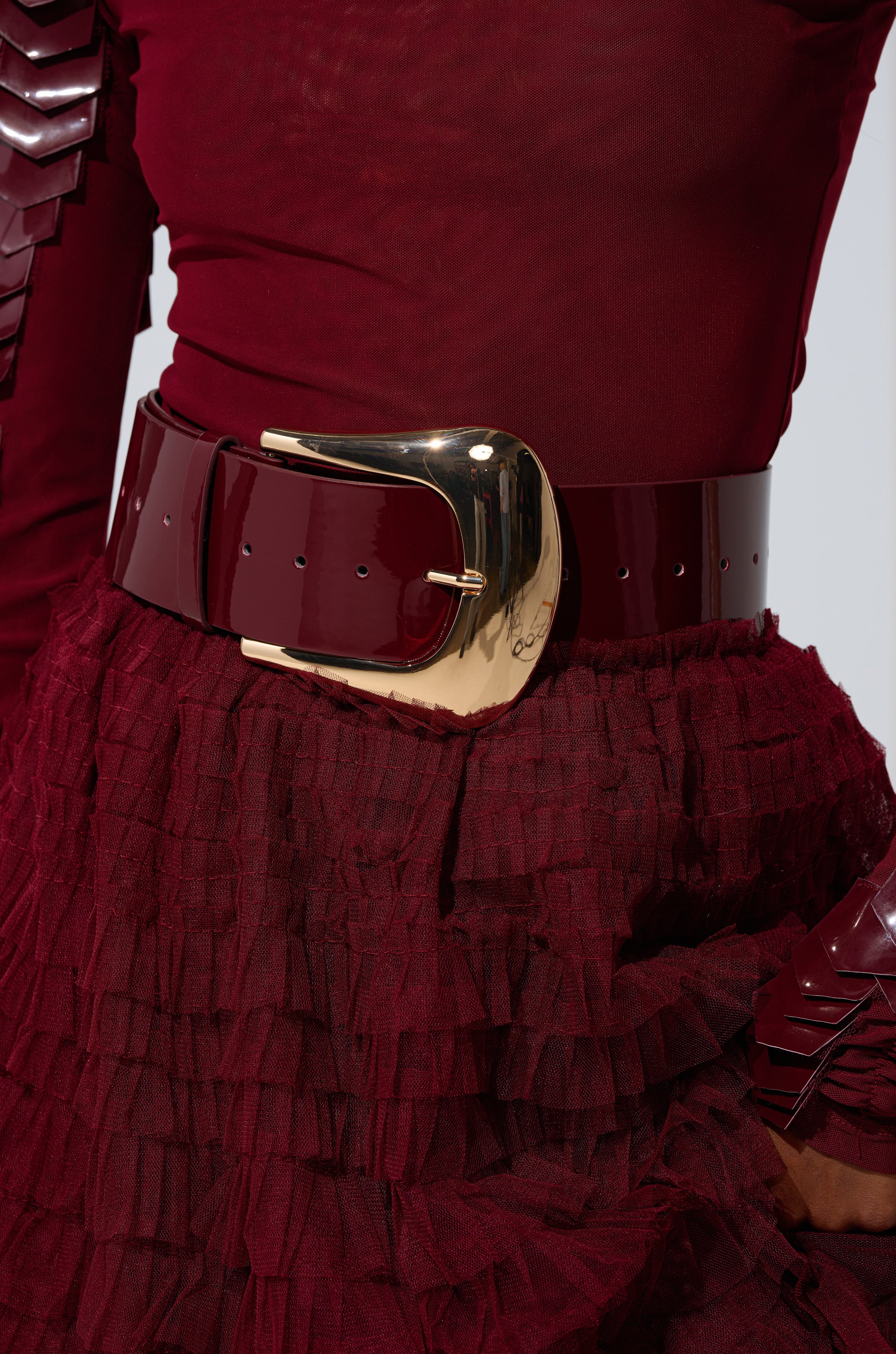 A person wearing a sheer burgundy top, a textured burgundy skirt, and the CINCH IT UP BELT featuring a wide faux patent leather band with an adjustable shiny gold buckle.
