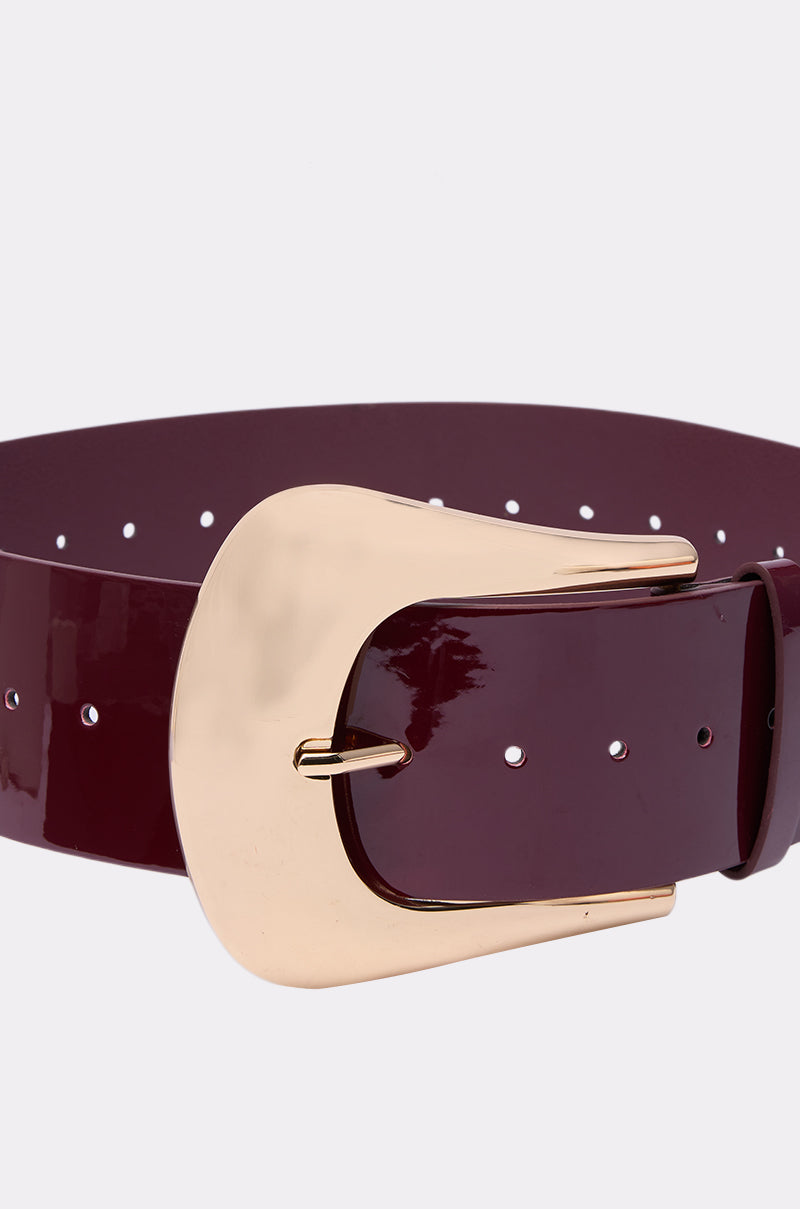 The CINCH IT UP BELT is a wide, glossy maroon faux patent leather belt with multiple adjustment holes and a large shiny gold buckle, offering style and versatility. Photographed against a plain white background.