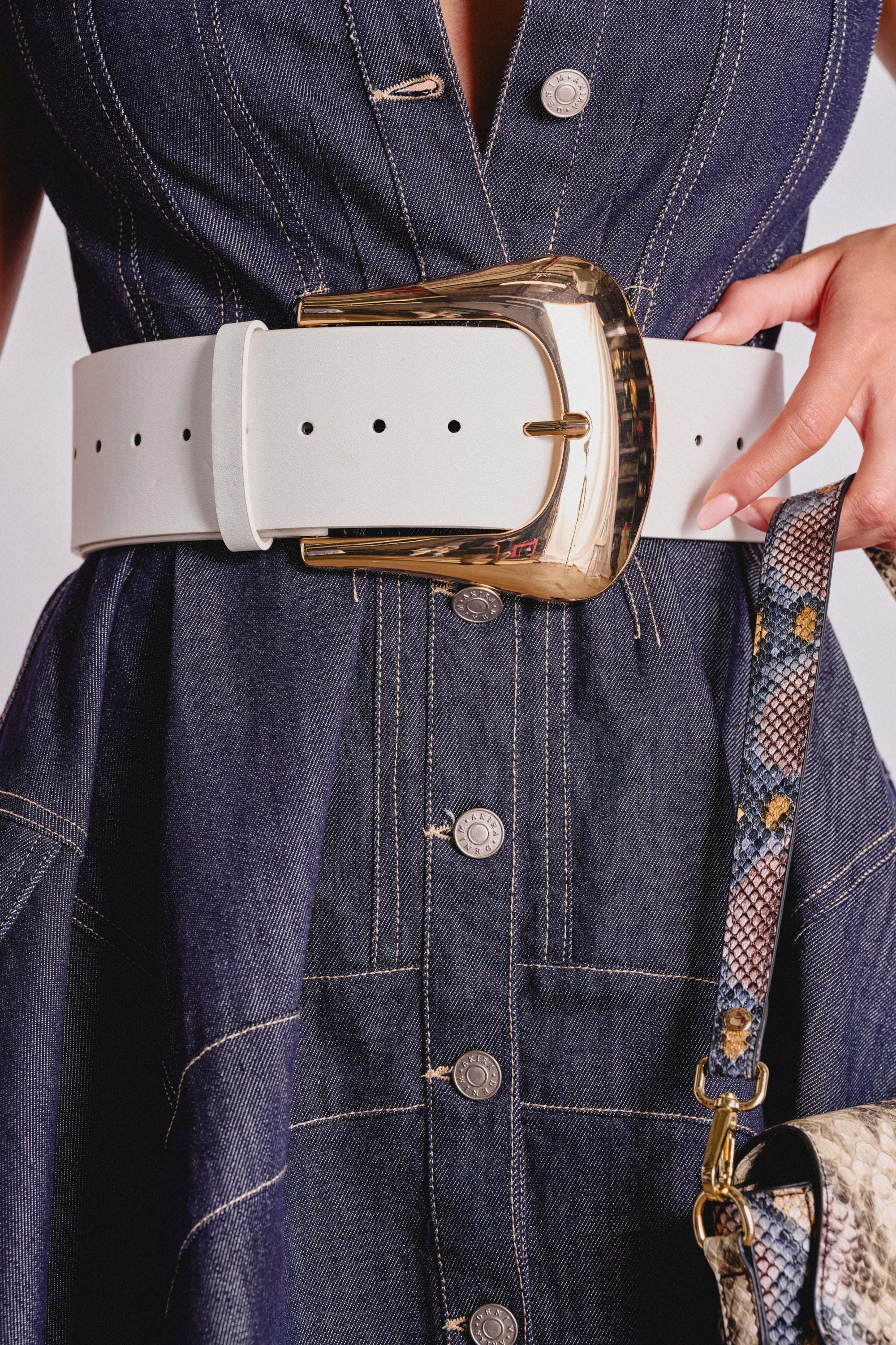 A close-up of a person wearing a denim dress with metal buttons, styled with the SIZE MATTERS OVERSIZED BUCKLE BELT in white faux leather, and holding a snakeskin-patterned handbag.