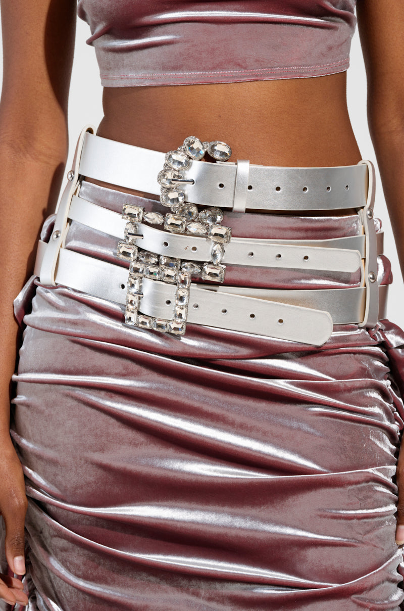 A person dresses in a shiny, ruched pink skirt and top, highlighted by the GET ON MY LEVEL MULTI BUCKLE BELT—an oversized metallic silver faux leather accessory adorned with multiple large rhinestone-encrusted buckles. The ensemble contrasts textures with its metallic sheen and sparkling details. 1