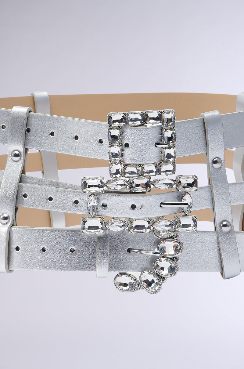 A detailed shot of the waist-cinching GET ON MY LEVEL MULTI BUCKLE BELT, showcasing its metallic silver faux leather design with multiple horizontal straps and a square buckle embellished with sparkling rhinestones, set against a light background. 4