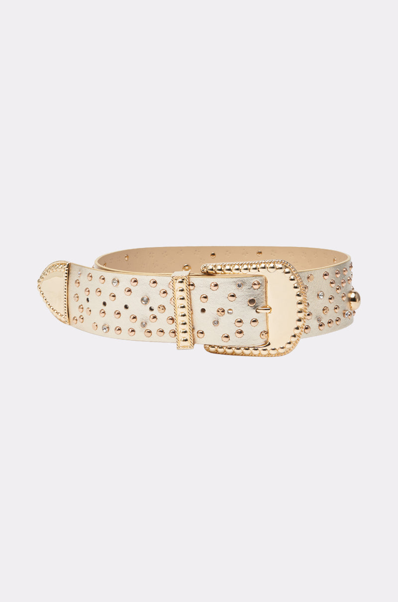 The DONNA TELL ME BELT is a beige leather belt featuring gold studs and a decorative, western-inspired gold buckle, displayed on a plain light background. 1