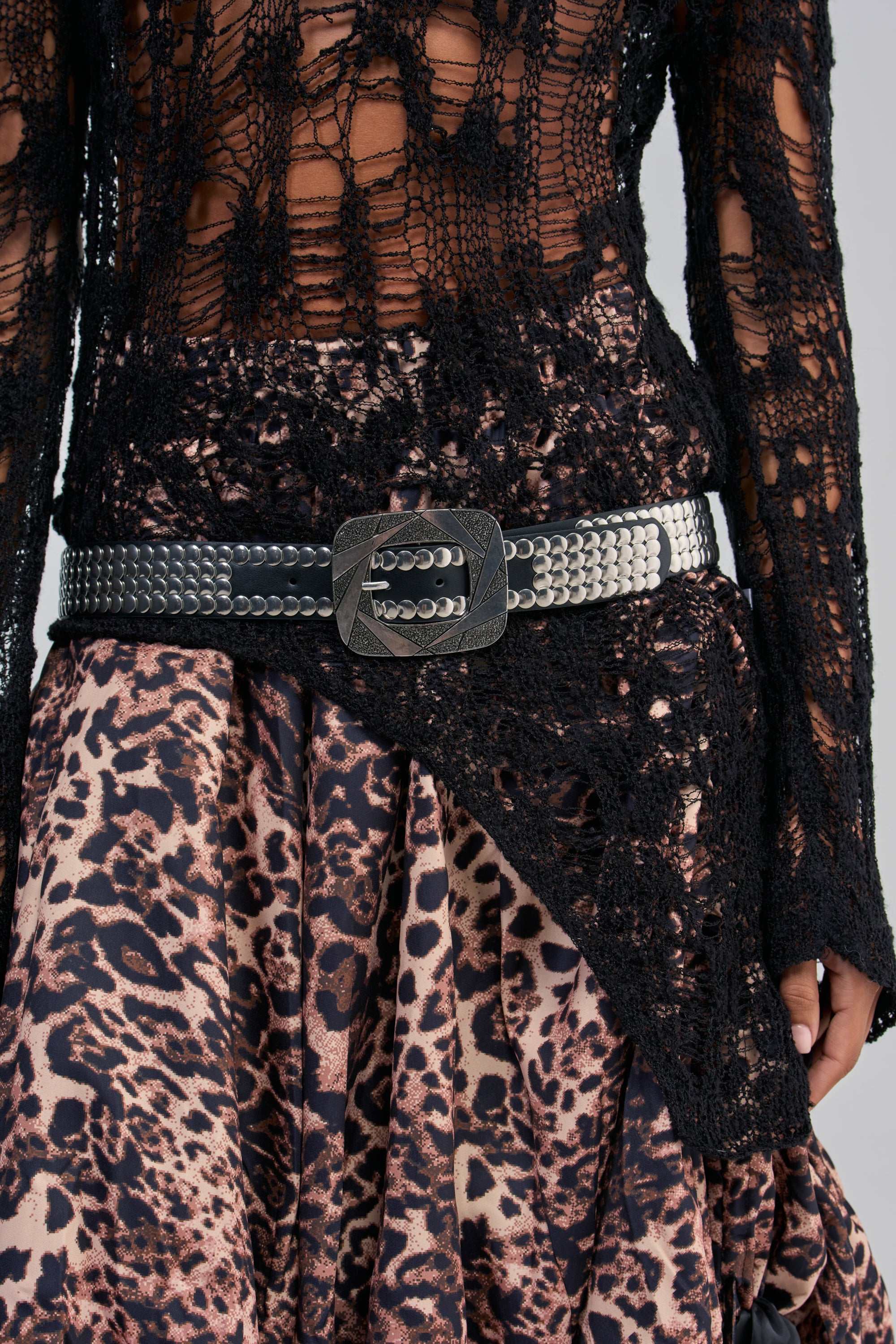 A person wears a black crochet top, the HEAVY METAL STUD BELT—a wide black faux leather belt with a large buckle—and a brown and black leopard print skirt.