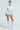 A model stands confidently against a light blue background, wearing a crocheted sweater with bows and SHAKE IT BABY RUFFLE SHORT IN BABY BLUE. She pairs the look with knee-high lace socks and white heels, right hand on her hip, gazing directly at the camera.