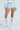 Adorned in the SHAKE IT BABY RUFFLE SHORT IN BABY BLUE, a crochet top, sheer white knee-high socks, and white high heels, the ensemble exudes playful and vintage charm with floral lace touches against a neutral backdrop.