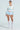 A model poses in a light blue crochet sweater with large bows over a matching top, white waistband, and SHAKE IT BABY RUFFLE SHORT IN BABY BLUE. She finishes the look with white lace knee-high socks and heels against a plain light blue background.