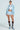 With glasses perched on their nose and hand on hip, a person poses stylishly against a plain backdrop, wearing a shiny light blue jacket, white shirt, Knee-high boots with intricate designs, and the SHAKE IT BABY RUFFLE SHORT IN BABY BLUE.