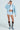 Wearing a light blue sequin jacket, white button-up shirt with a cross necklace, STYLE IT BABY RUFFLE SHORTS in baby blue, and brown embellished high-heeled boots, they complete the look with sunglasses and stylishly pulled-back hair.