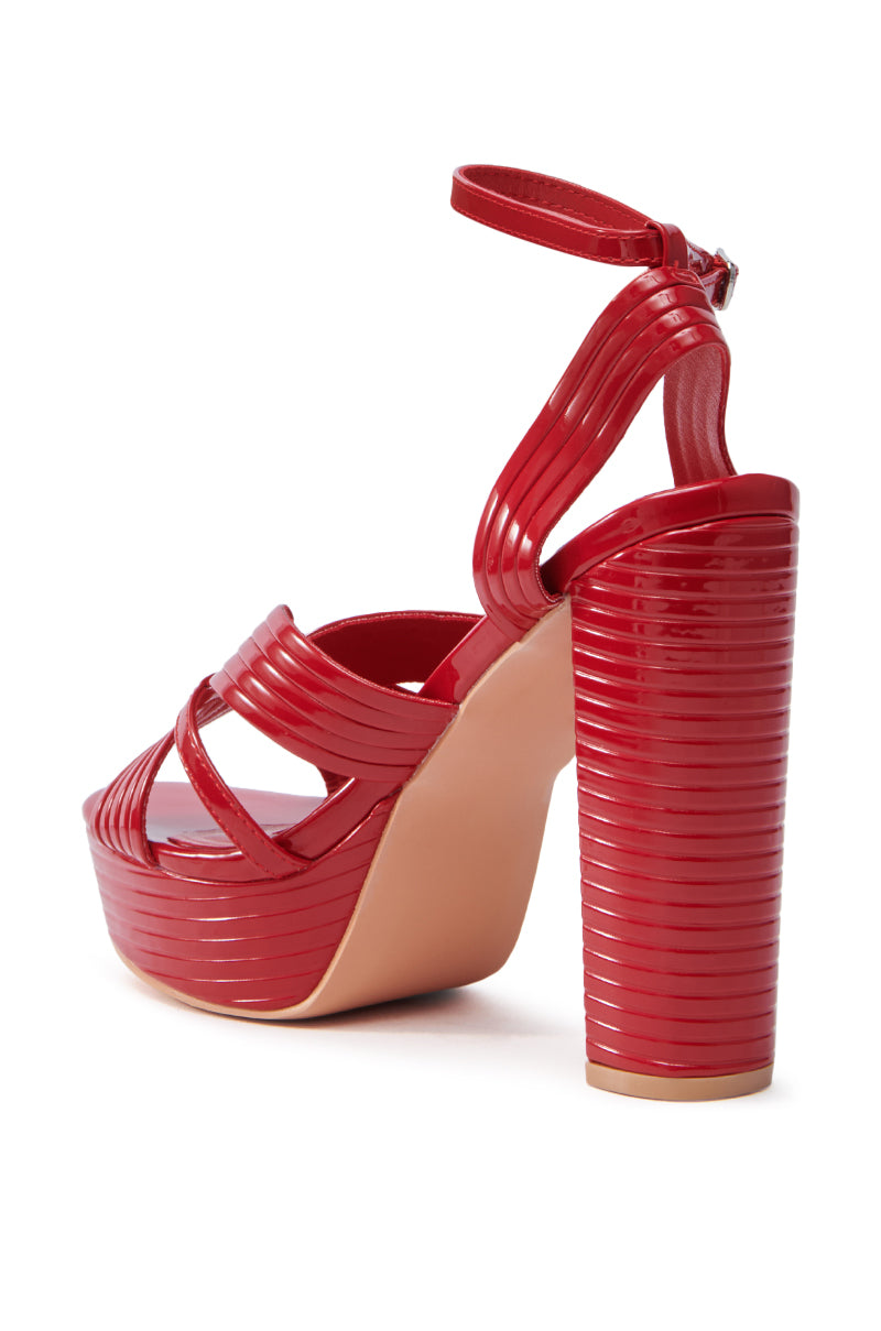 The AZALEA WANG RED STRAPPY PLATFORM SANDAL features a glossy red faux leather design with crisscross straps, a chunky heel, and a platform sole for added height. An ankle strap with a buckle provides a secure fit.