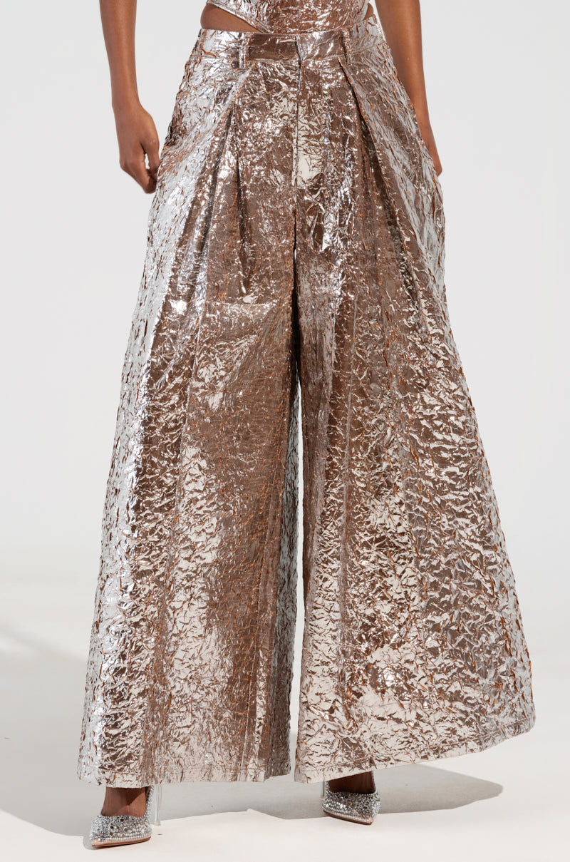 A person wearing the STAR PANT, characterized by its shiny, crinkled metallic texture and wide-leg design, stands with hands in pockets. The high-rise style beautifully reflects light. The embellished shoes are visible at the bottom against a plain, neutral backdrop. 1