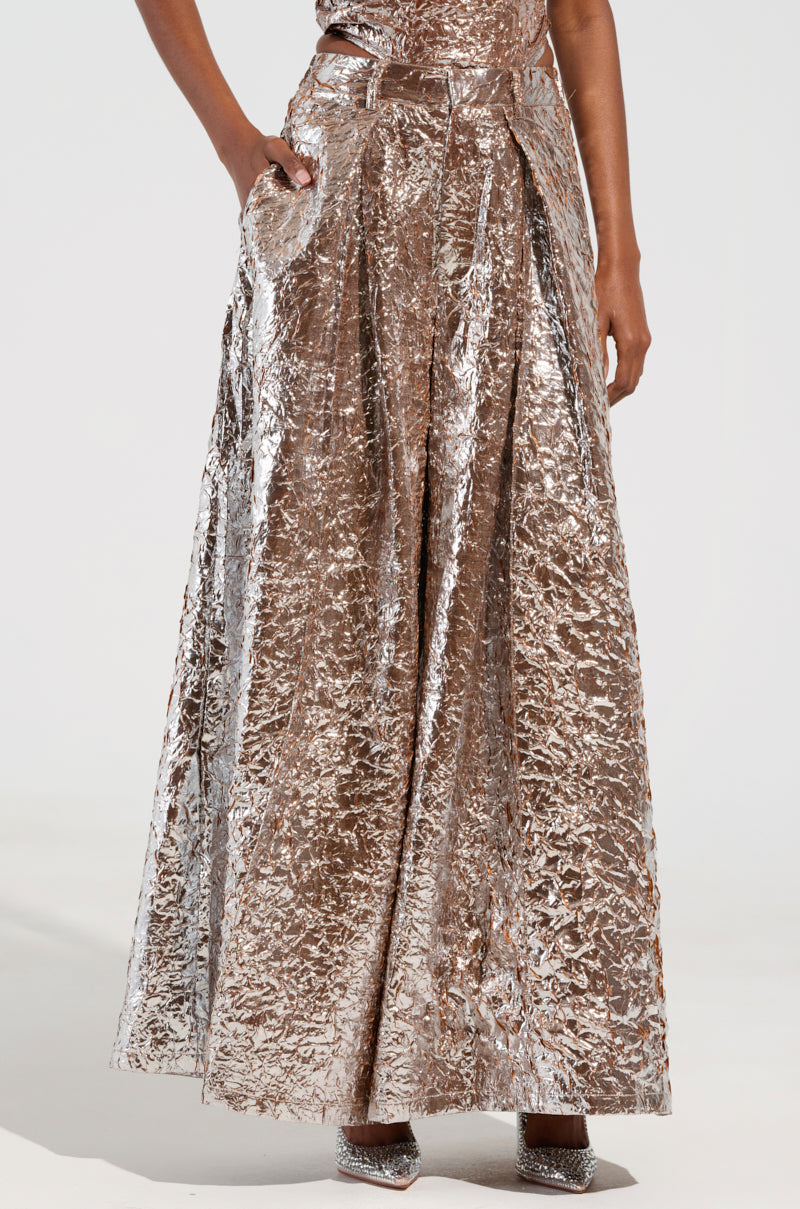 The person is dressed in the STAR PANT, featuring a shiny, metallic finish with a wide-leg silhouette and high-rise design, standing with one hand in a pocket. The ensemble is completed with sparkling heels, causing the fabric to reflect the light beautifully. 2