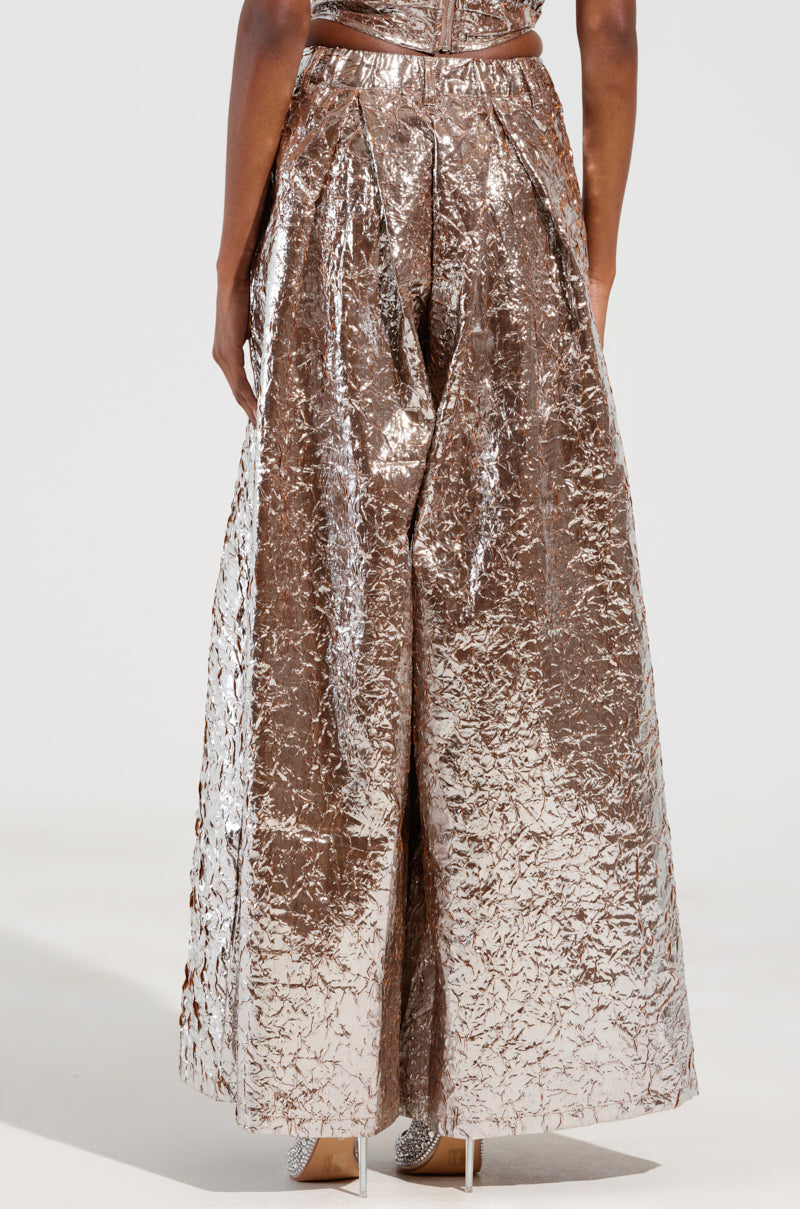 A person is sporting the STAR PANT, which features high-rise, metallic bronze fabric with a crinkled finish and wide-leg silhouette. The garment shines brilliantly against the plain, light backdrop, capturing attention with its shimmering texture. Only the lower half of the body is visible in the frame. 4