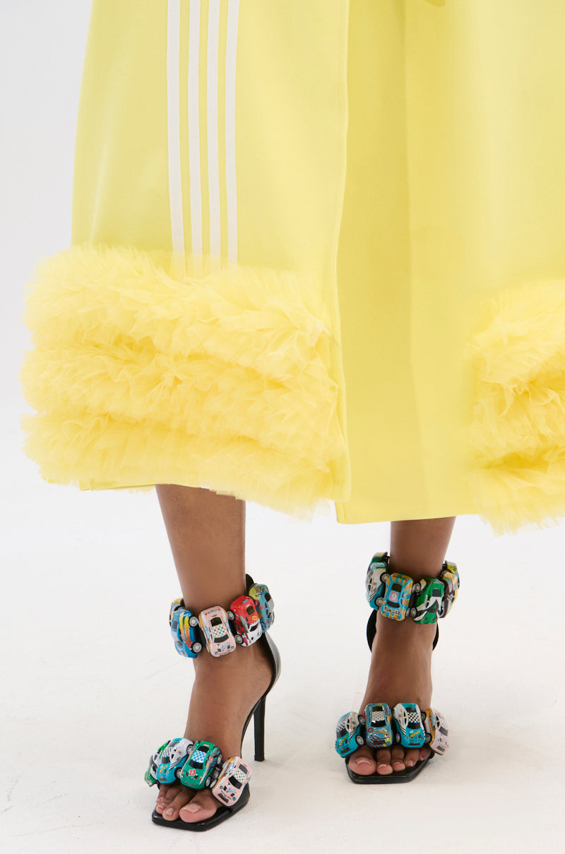 A person in a yellow ruffled skirt pairs it with AZALEA WANG STARLITE BLACK CARS STILETTO SANDALS featuring an open square toe and miniature toy car accents on the ankle and toe straps.