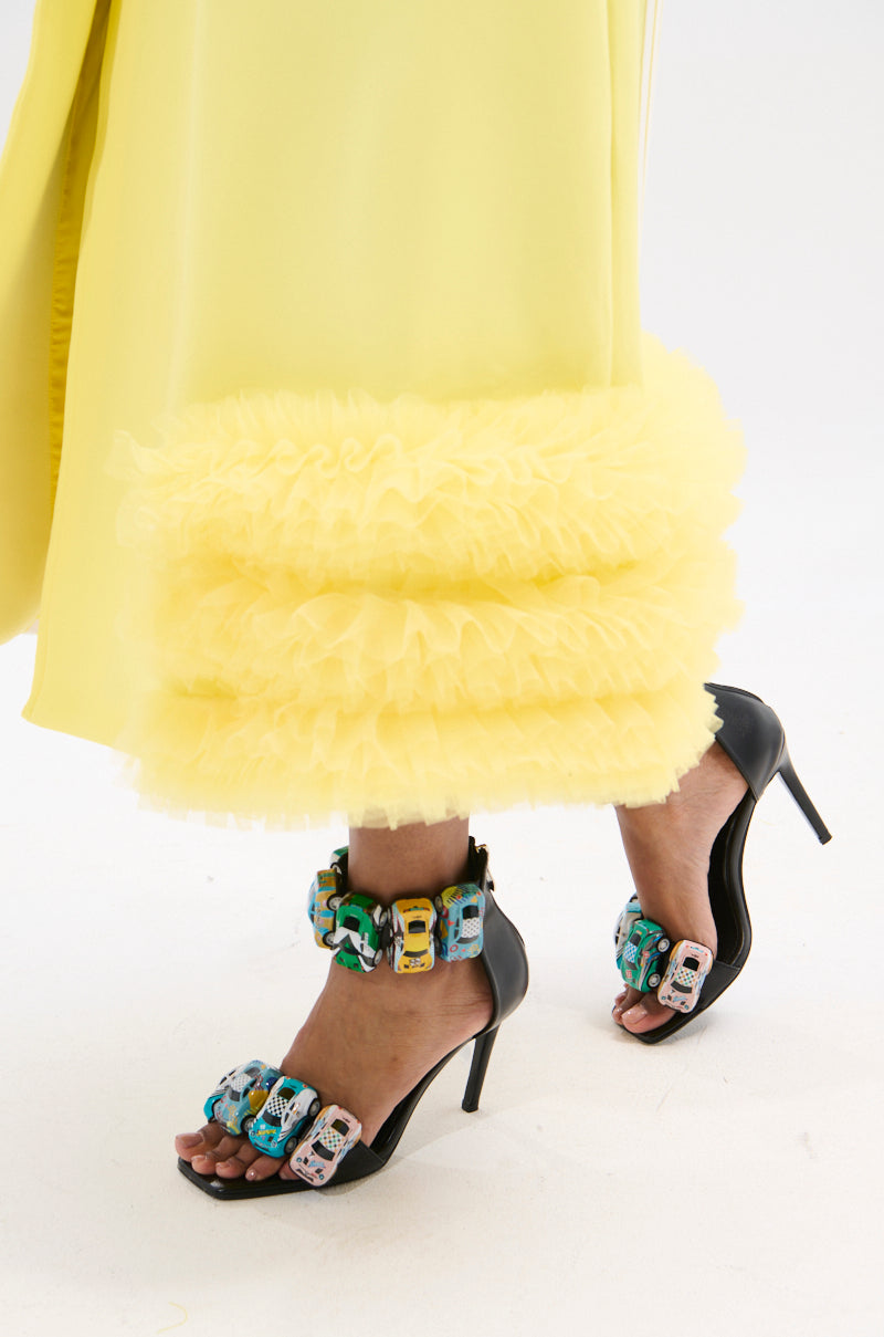 A person wears yellow ruffled pants and AZALEA WANG STARLITE BLACK CARS STILETTO SANDALS with colorful toy car accents, their open square toes highlighted against a white background.