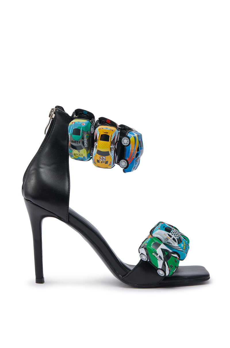 The AZALEA WANG STARLITE BLACK CARS STILETTO SANDAL is a black faux leather sandal with an open square toe, colorful toy car straps in green, yellow, blue, and black, a back zipper, and a playful modern design.