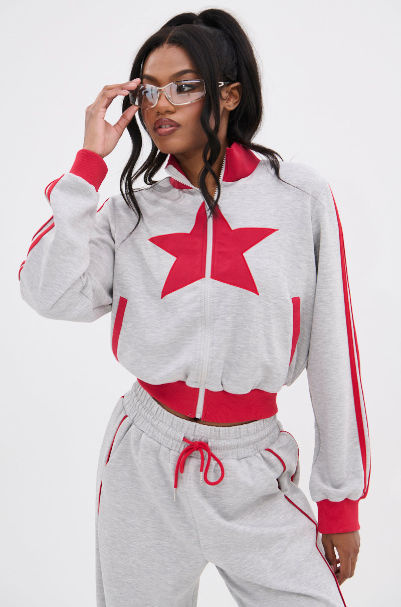 A woman in clear glasses wears the ALL STAR FULL ZIP MOCK NECK SWEATSHIRT, featuring red trim and a bold star appliqué, as she stands against a white background and holds her glasses with one hand.