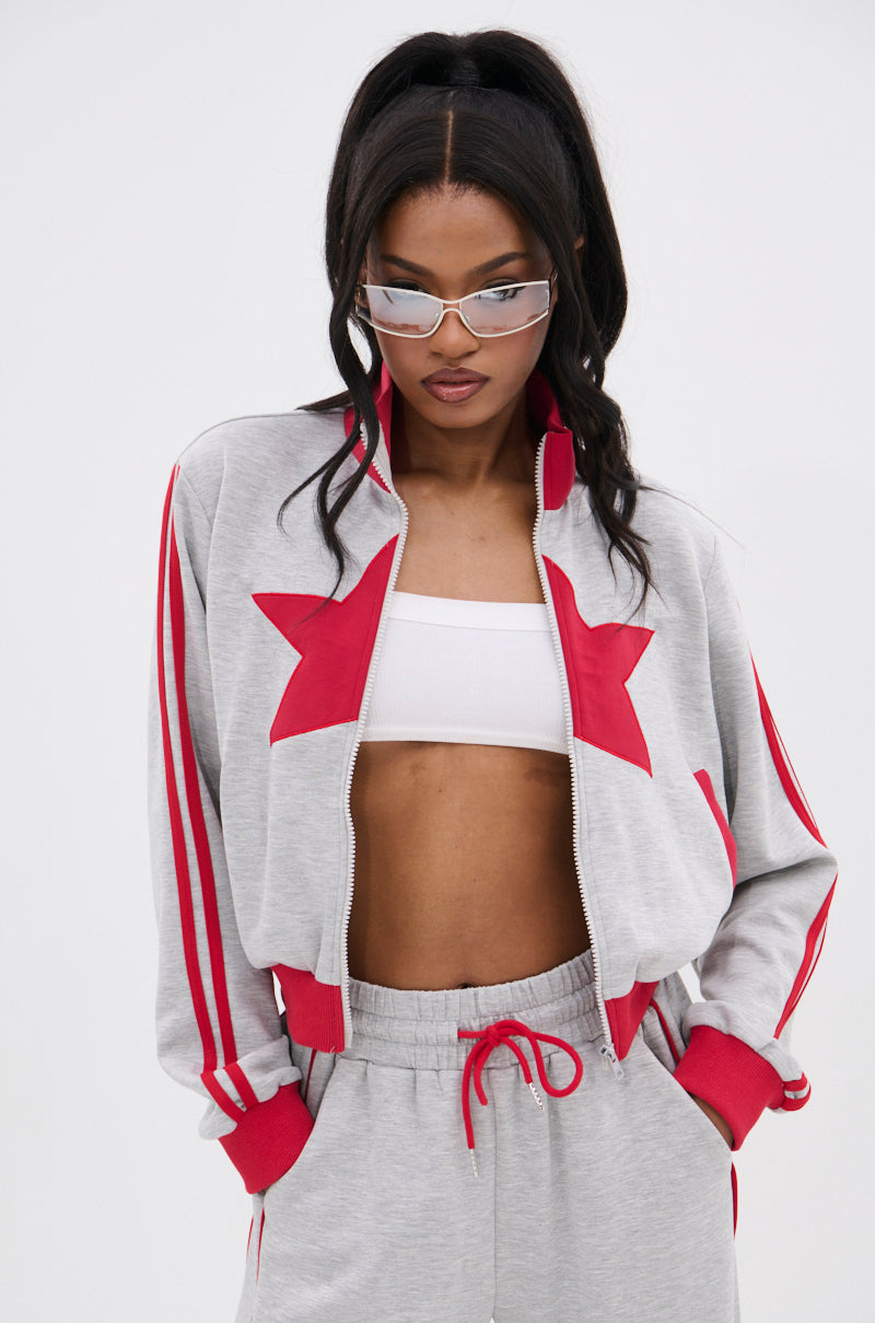 A woman stands hands-in-pockets against a white background wearing the ALL STAR FULL ZIP MOCK NECK SWEATSHIRT with star appliqué, a white crop top, silver sunglasses, and her hair in a high ponytail.
