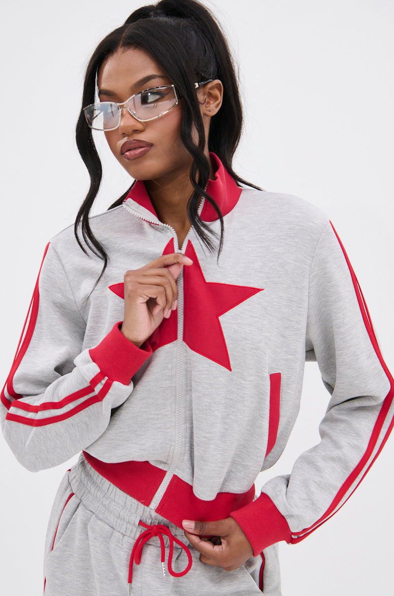 A woman in clear glasses and the ALL STAR FULL ZIP MOCK NECK SWEATSHIRT with a bold red star on the chest stands against a plain white background, holding her jacket’s zipper.