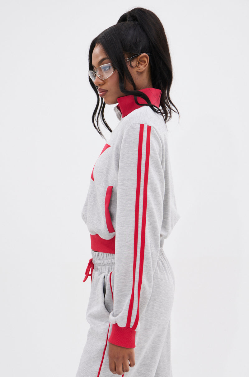 A woman in profile wears the ALL STAR FULL ZIP MOCK NECK SWEATSHIRT, a light gray zip-up with red stripes and star appliqué, styled with clear glasses and a high ponytail, posed against a plain white background.