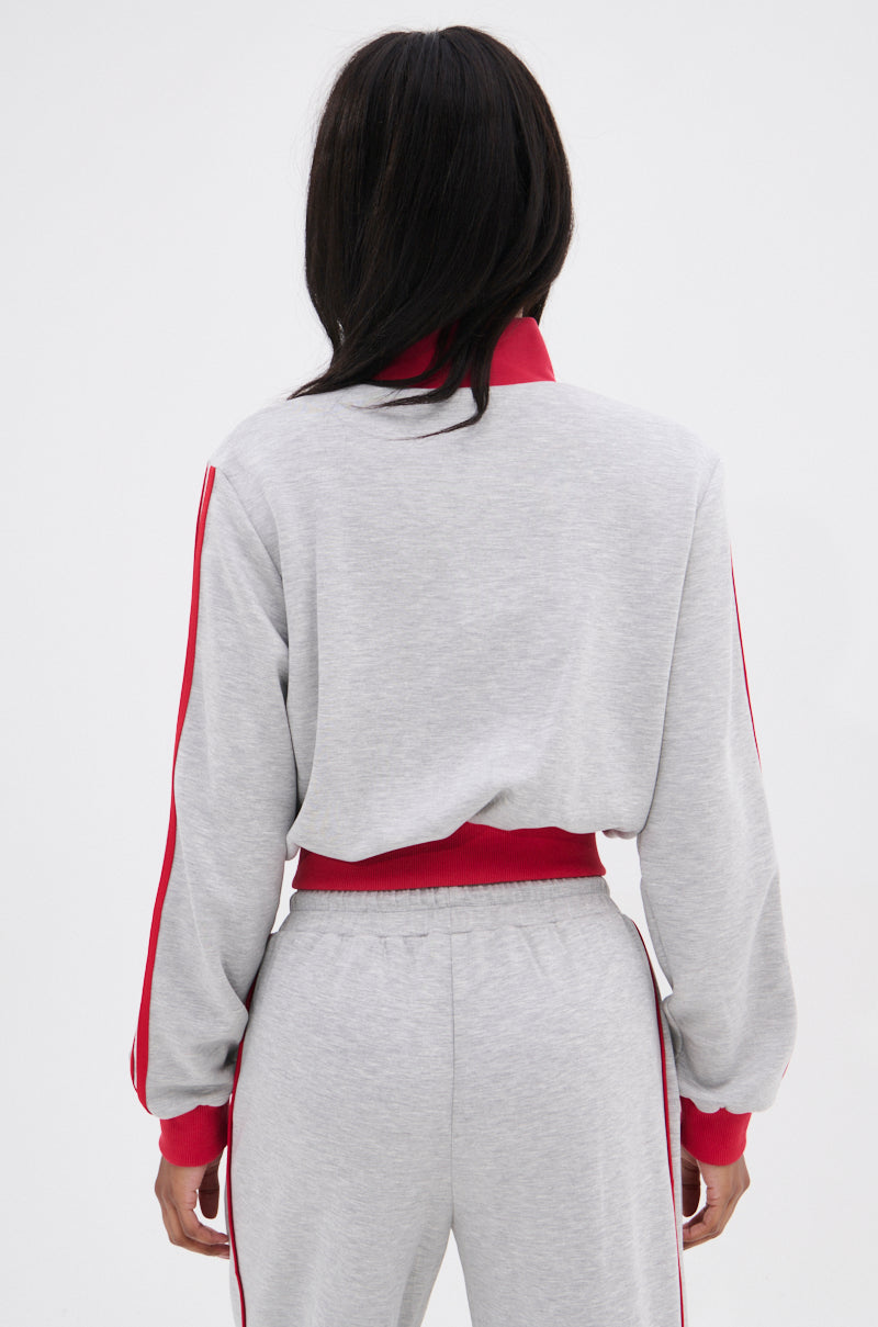 A person with long, dark hair is seen from behind wearing the ALL STAR FULL ZIP MOCK NECK SWEATSHIRT in light gray, featuring red accents and star appliqué on the collar, cuffs, waistband, and down the sleeves.