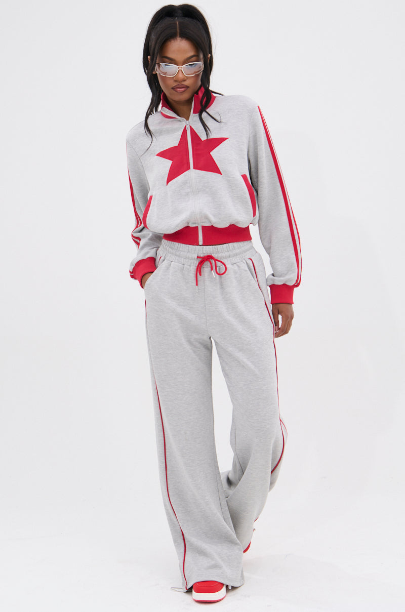 A woman with long dark hair and clear glasses wears the ALL STAR FULL ZIP MOCK NECK SWEATSHIRT in light gray, accented with a red star appliqué, red sleeve stripes, matching pants, and red shoes.