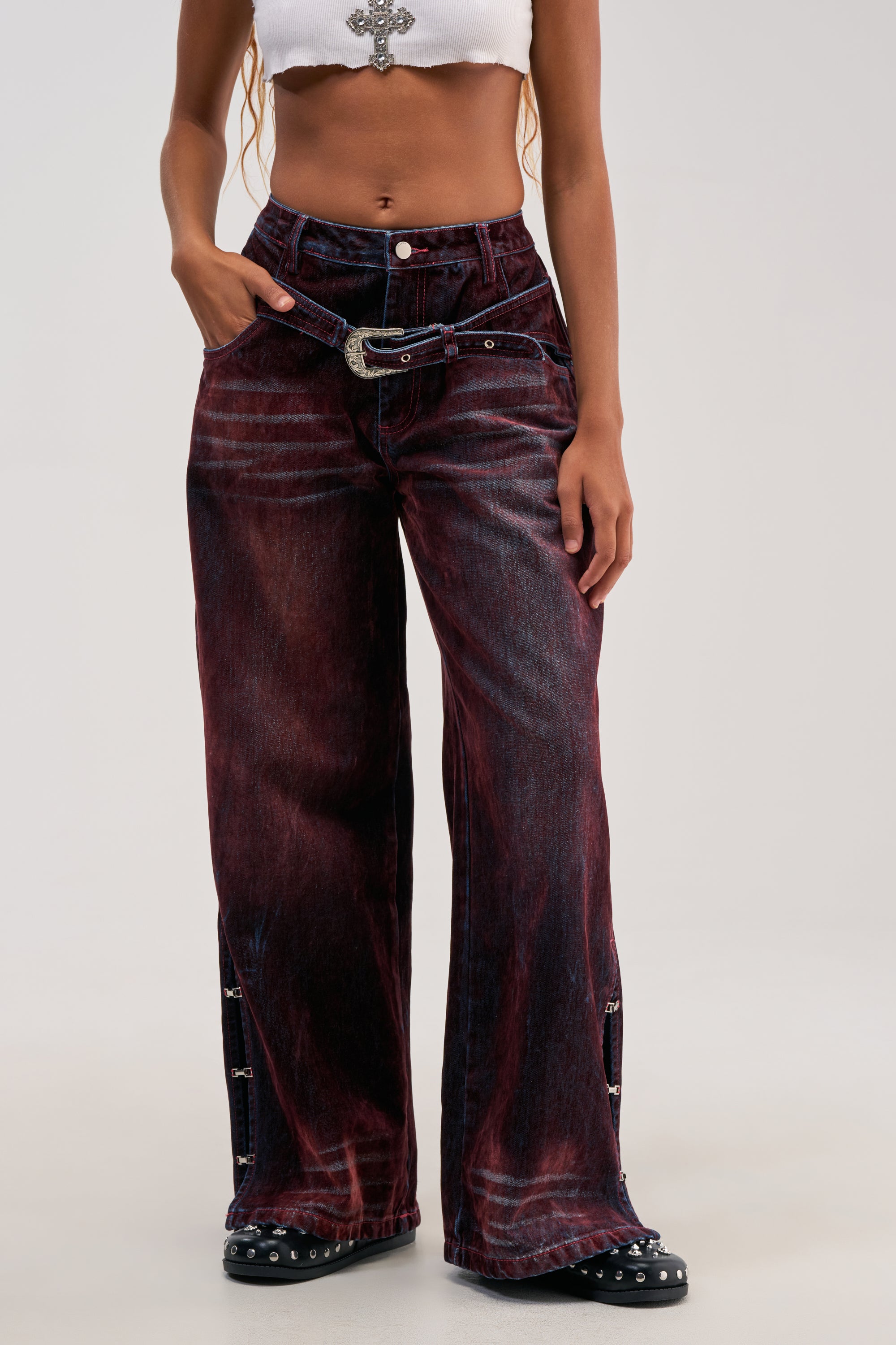 A woman wears a white crop top and the MIDNIGHT MERLOT VELVET DENIM PANTS—western-inspired, wide-leg jeans in burgundy velvet—styled with a decorative belt and black studded shoes. Her right hand is tucked into her pocket, set against a plain, light background.