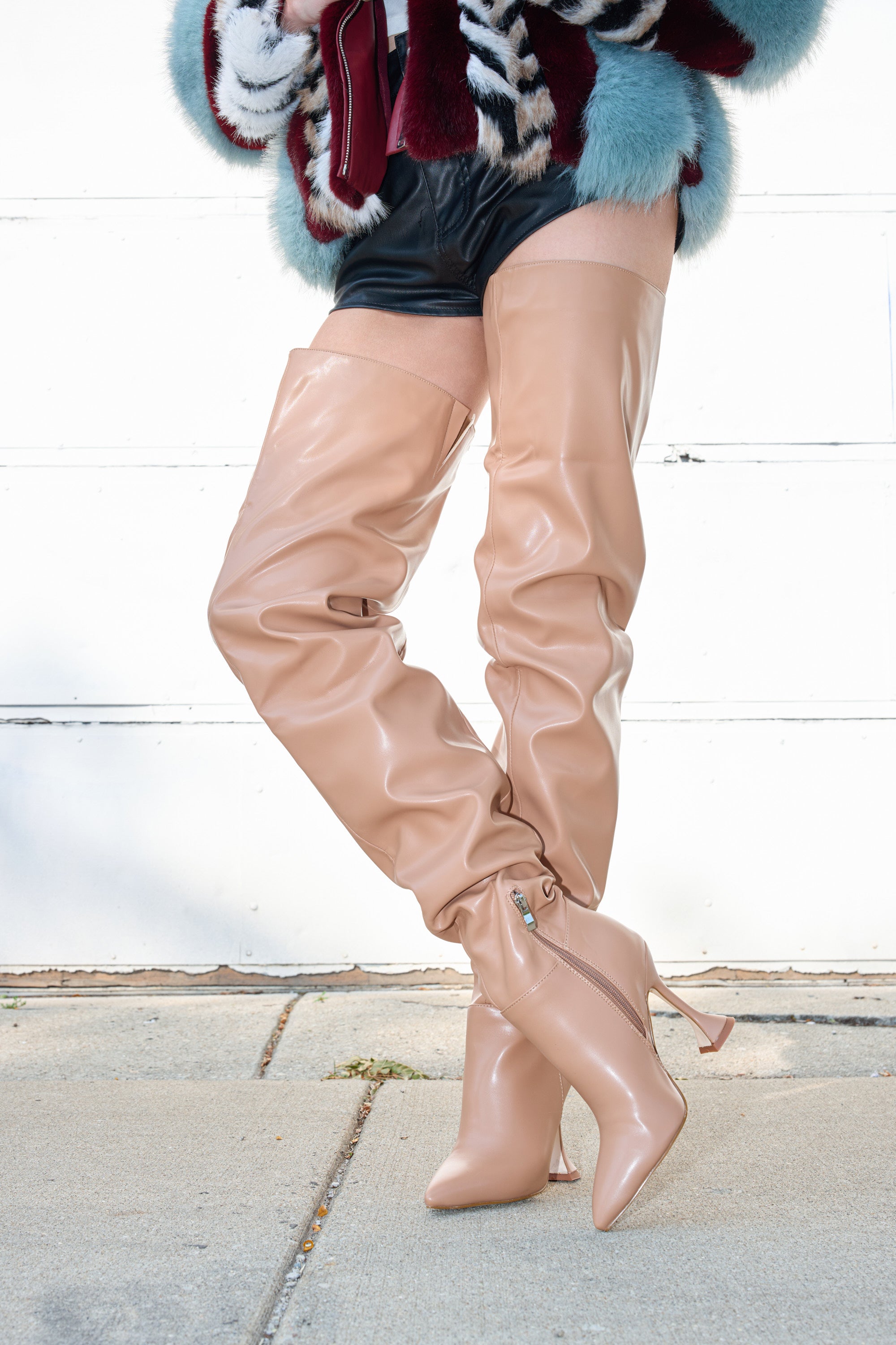 A person in AZALEA WANG STEPHAN BEIGE PU FLARED HEEL RUCHED BOOTS, black shorts, and a colorful fur coat stands on the sidewalk with legs crossed at the ankles. The backdrop is a white wall.