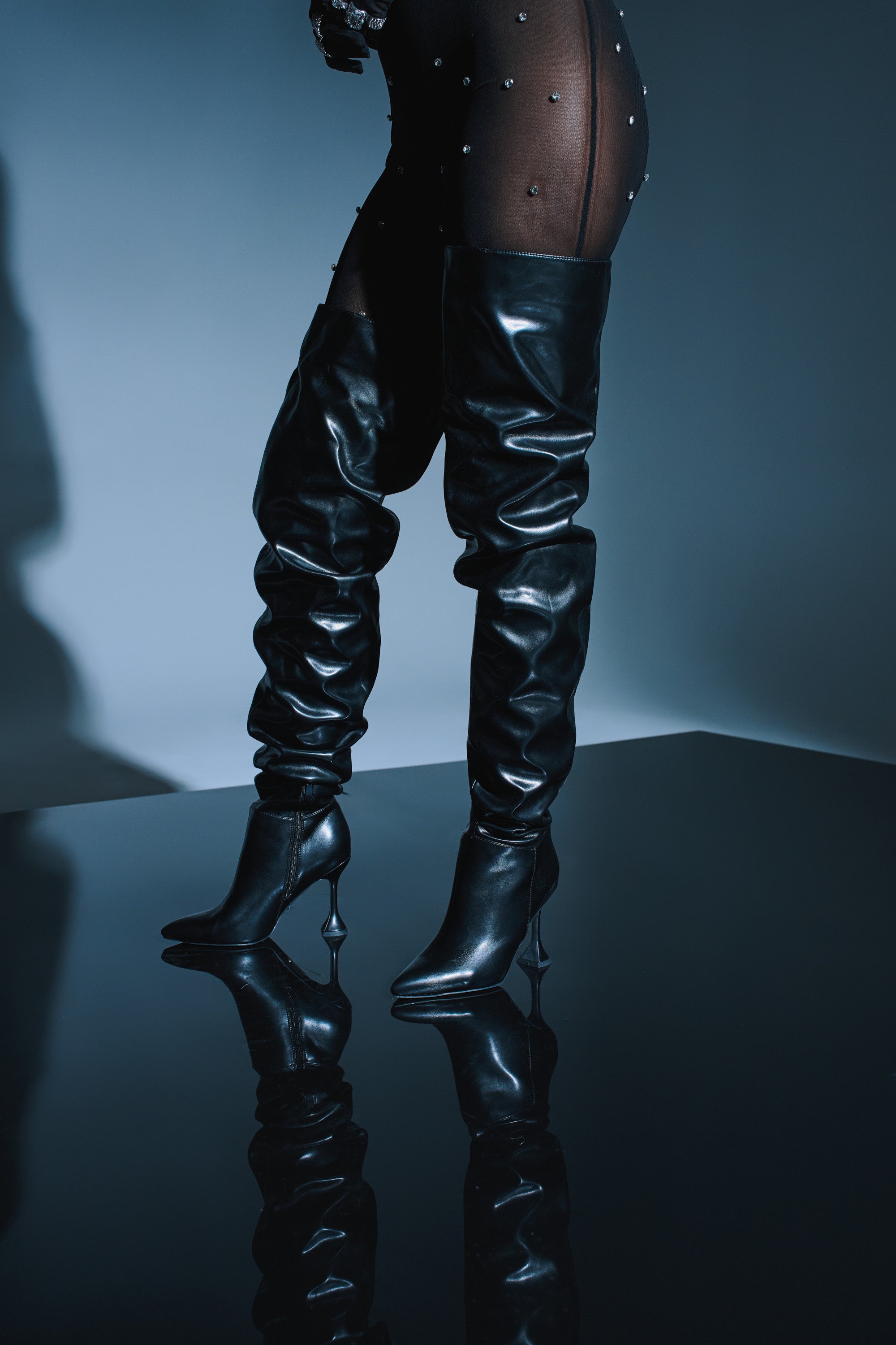 Wearing sheer black tights with small embellishments and the AZALEA WANG STEPHAN BLACK BOOT, a person stands on a reflective surface, creating a stylish and moody scene.