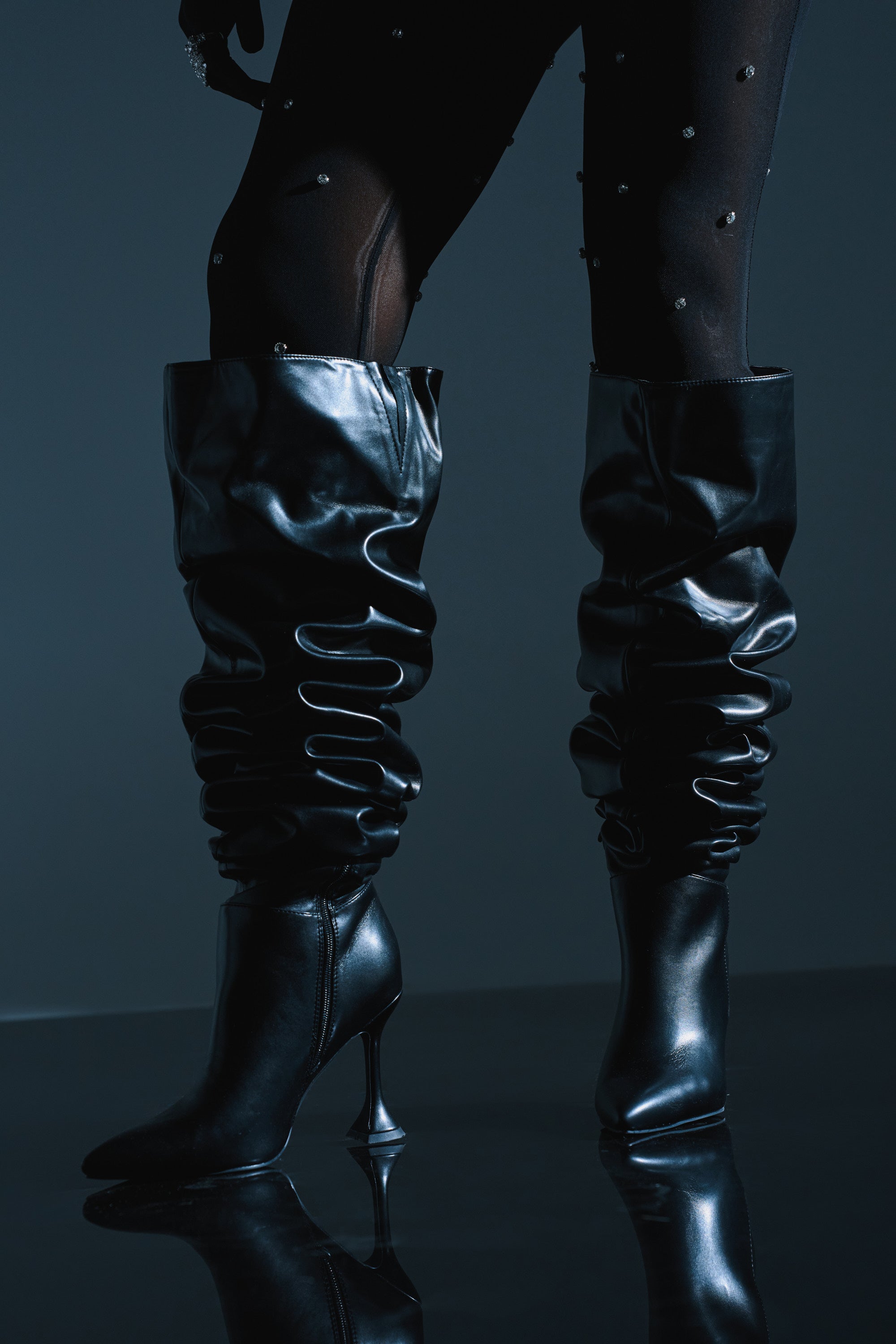A person wears the AZALEA WANG STEPHAN BLACK BOOT—black, slouchy faux leather stilettos—with sheer black embellished tights, standing on a glossy dark surface against a dark background.