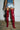 A person stands against a white brick wall, wearing AZALEA WANG STEPHAN BURGUNDY BOOT thigh-highs, distressed denim shorts with a tartan lining, and a blue plaid top. They also carry a black handbag in their left hand. The floor is a rough, unfinished surface.