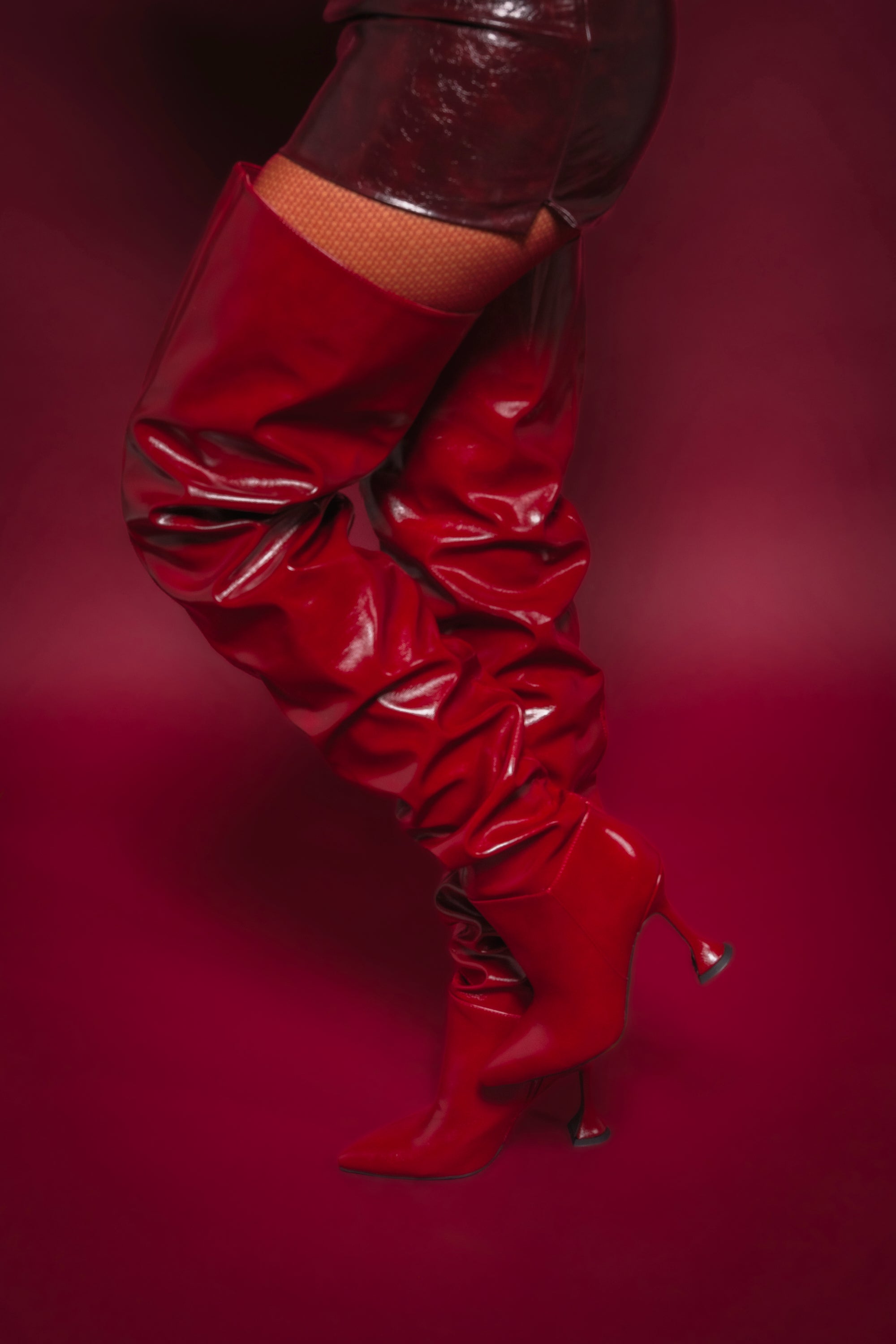 A person wearing AZALEA WANG STEPHAN BURGUNDY BOOTS and red shorts poses against a red background, with one leg slightly bent and the other extended. 0