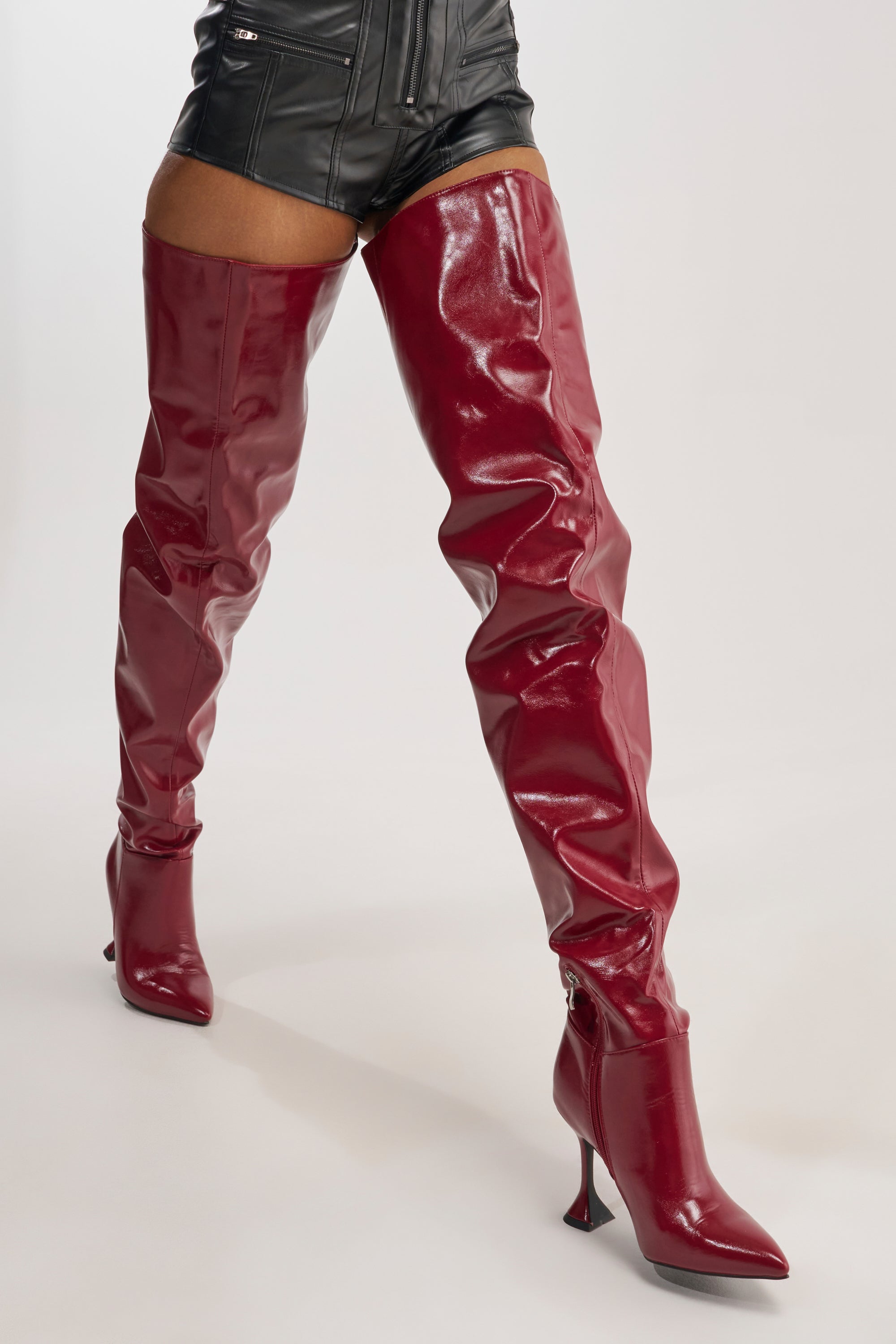 A person is wearing the AZALEA WANG STEPHAN BURGUNDY BOOT, featuring shiny, burgundy thigh-highs with stiletto heels, paired with black leather shorts and standing against a plain white background.