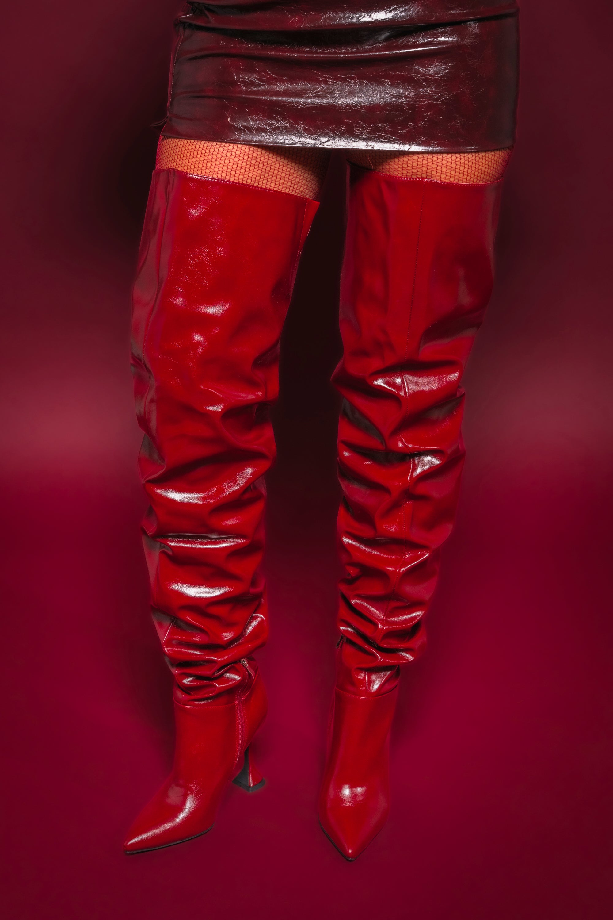 A person wearing the AZALEA WANG STEPHAN BURGUNDY BOOT with stiletto heels and pointed toes stands against a deep red background, paired with a glossy red mini skirt. 1