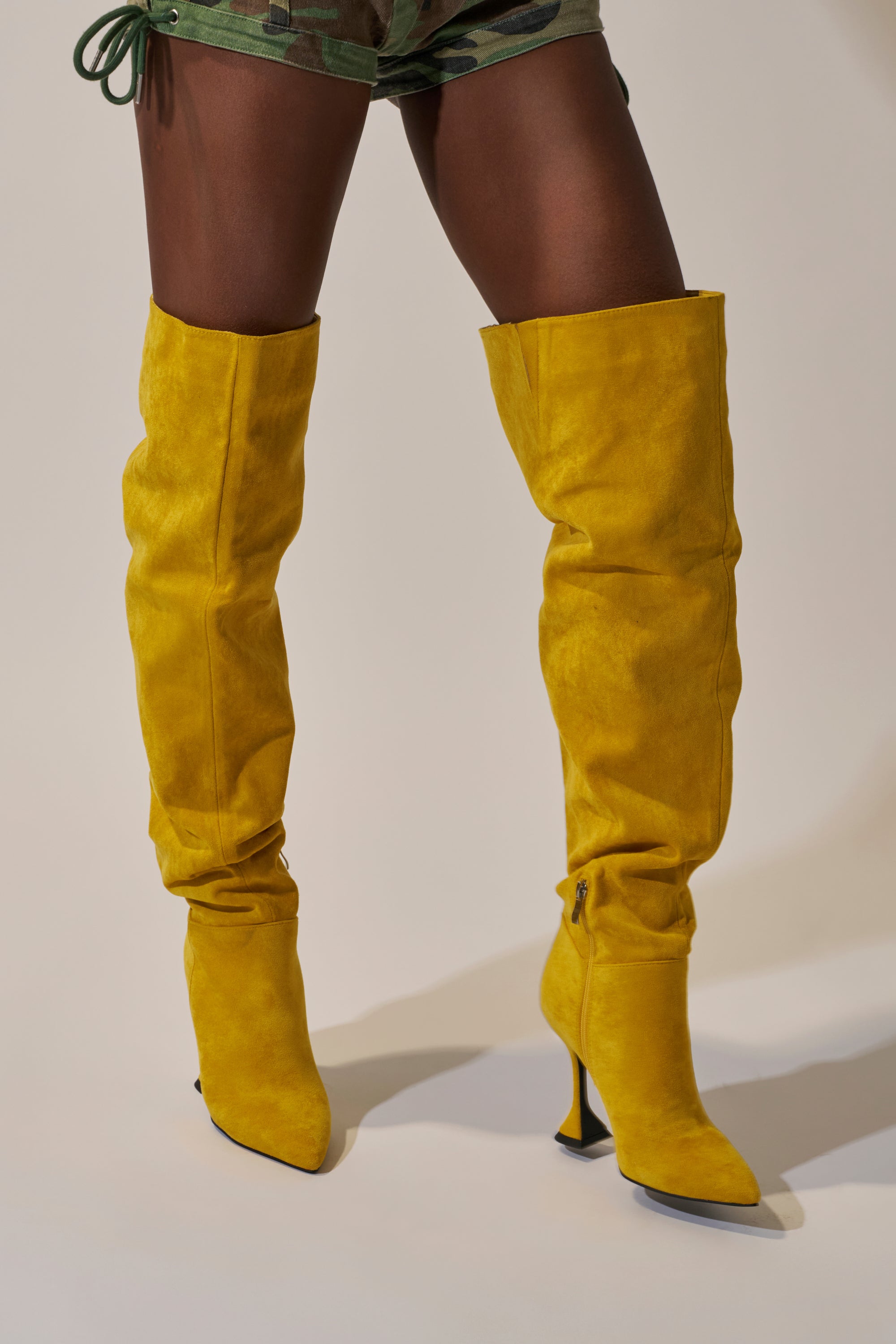 A person in camo shorts wears bold AZALEA WANG STEPHAN YELLOW SUEDE CHUNKY BOOTS, which reach above the knee and stand out against a neutral background.