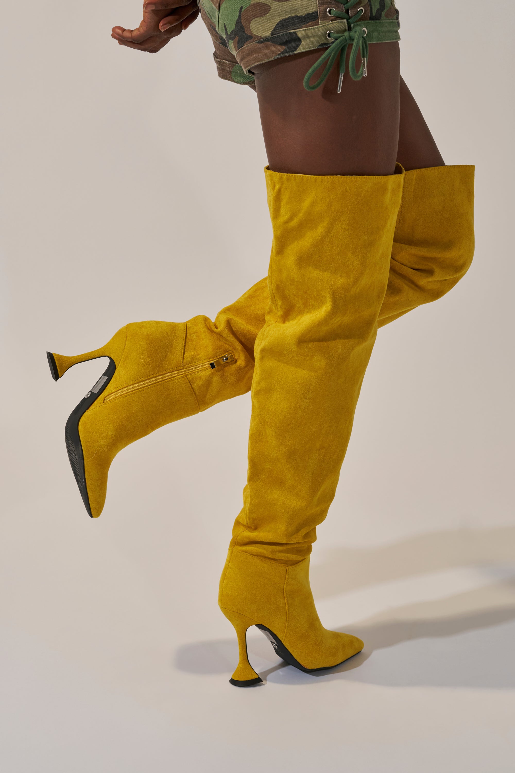 A person models the AZALEA WANG STEPHAN YELLOW SUEDE CHUNKY BOOT with green camouflage shorts, posing with one leg bent and the other straight against a neutral background.