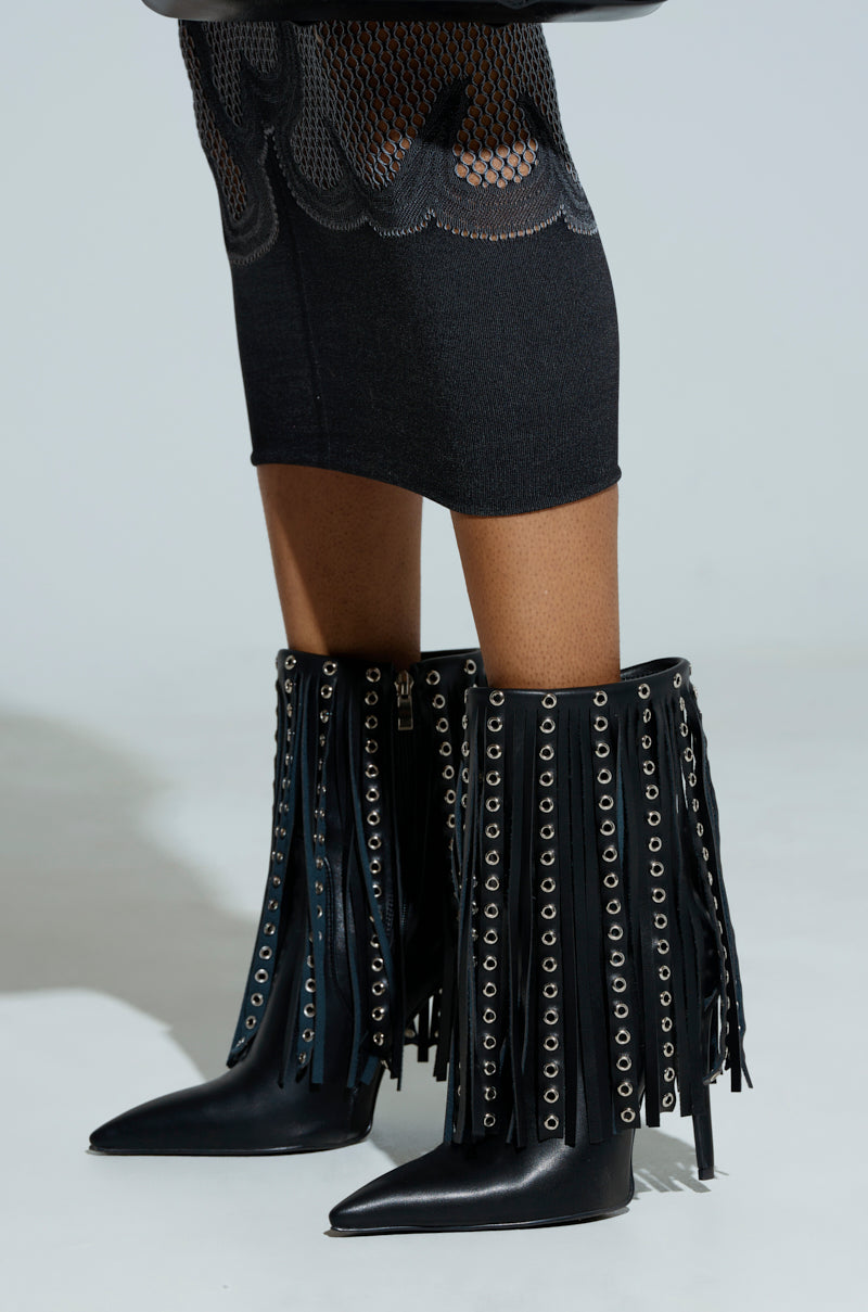 A person wearing a black skirt with mesh detailing and AZALEA WANG STERLING BLACK FRINGE STILETTO BOOTIE, adorned with metal eyelets. These pointed toe boots feature a high shaft, making a bold fashion statement.