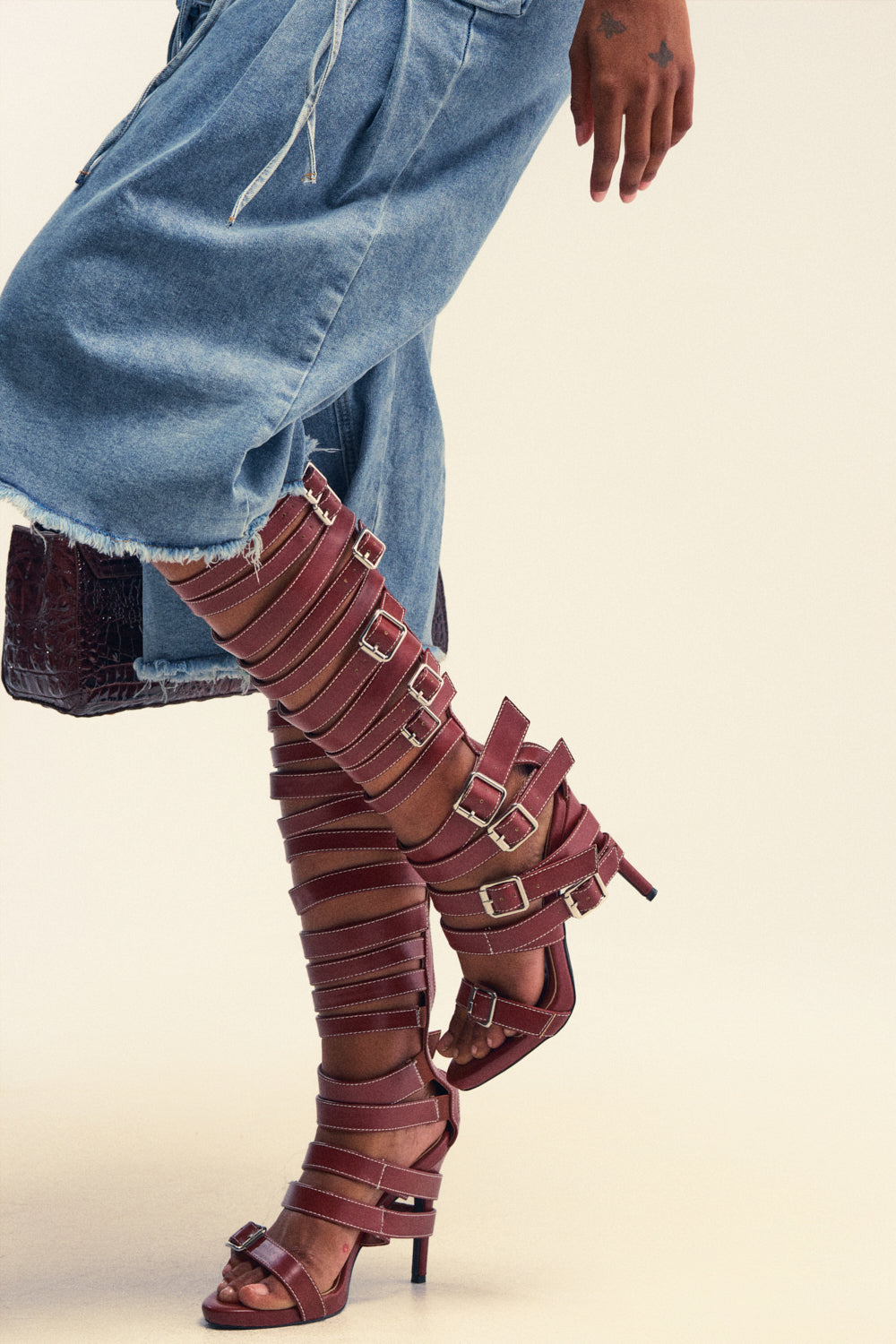 Wearing a denim skirt, the person sports AZALEA WANG STITCH BROWN PU HARDWARE EMBELLISHED STILETTO SANDALS—tall, strappy, with multiple buckles—and has a star tattoo on their hand.