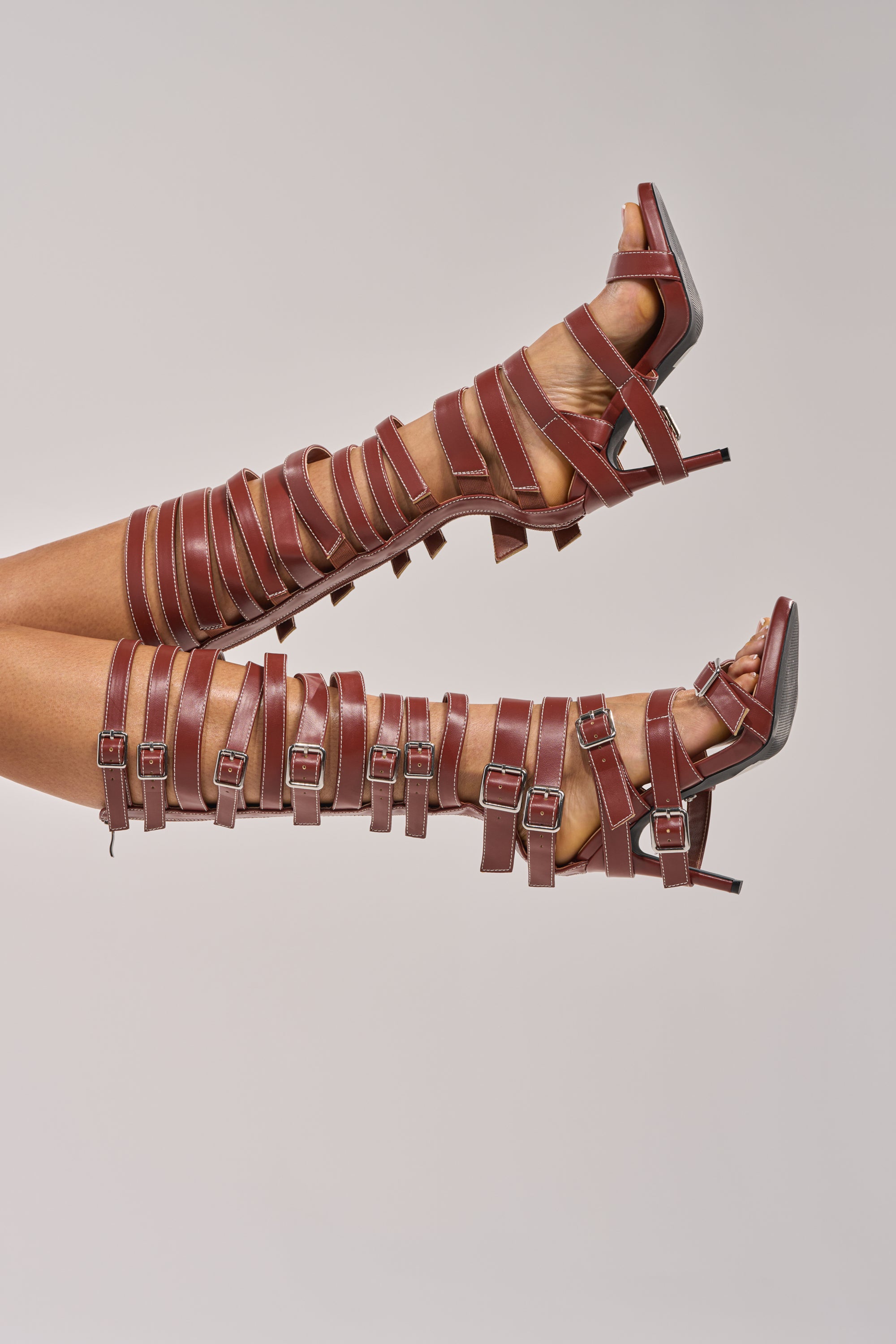 A pair of legs model the AZALEA WANG STITCH BROWN PU HARDWARE EMBELLISHED STILETTO SANDAL, featuring knee-high brown faux leather with multiple silver buckles, set against a plain light background.