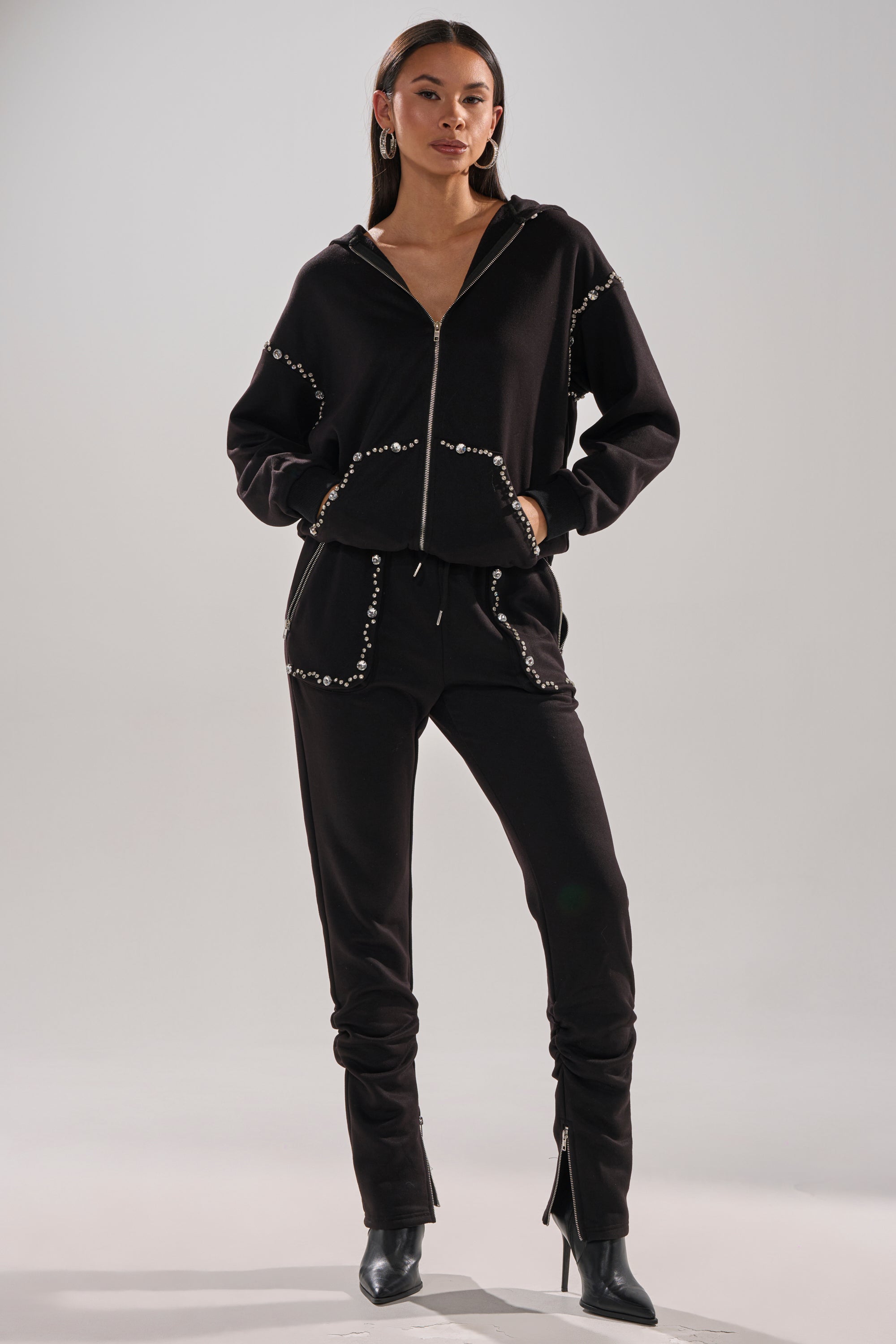 A woman wears the COZY HOLIDAY RHINESTONE JOGGER IN BLACK—featuring a black zip-up hoodie and matching joggers with white rhinestone trim—paired with black heeled boots, hands in pockets, facing forward against a plain background.