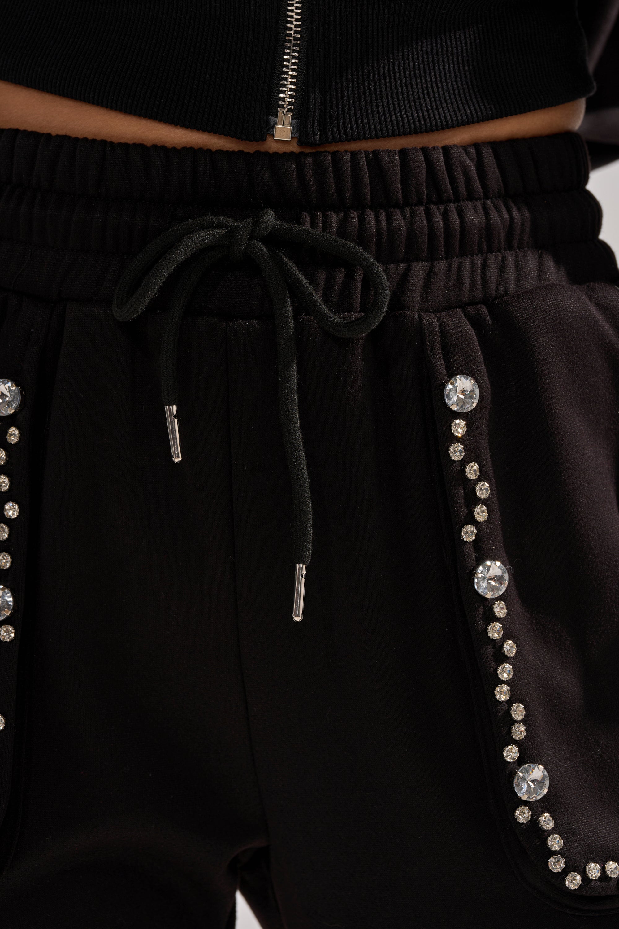 The COZY HOLIDAY RHINESTONE JOGGER IN BLACK features a drawstring waist and rows of sparkling small and large rhinestones along the pocket edges. A black zippered top is partially shown above the waistband, adding extra holiday flair.