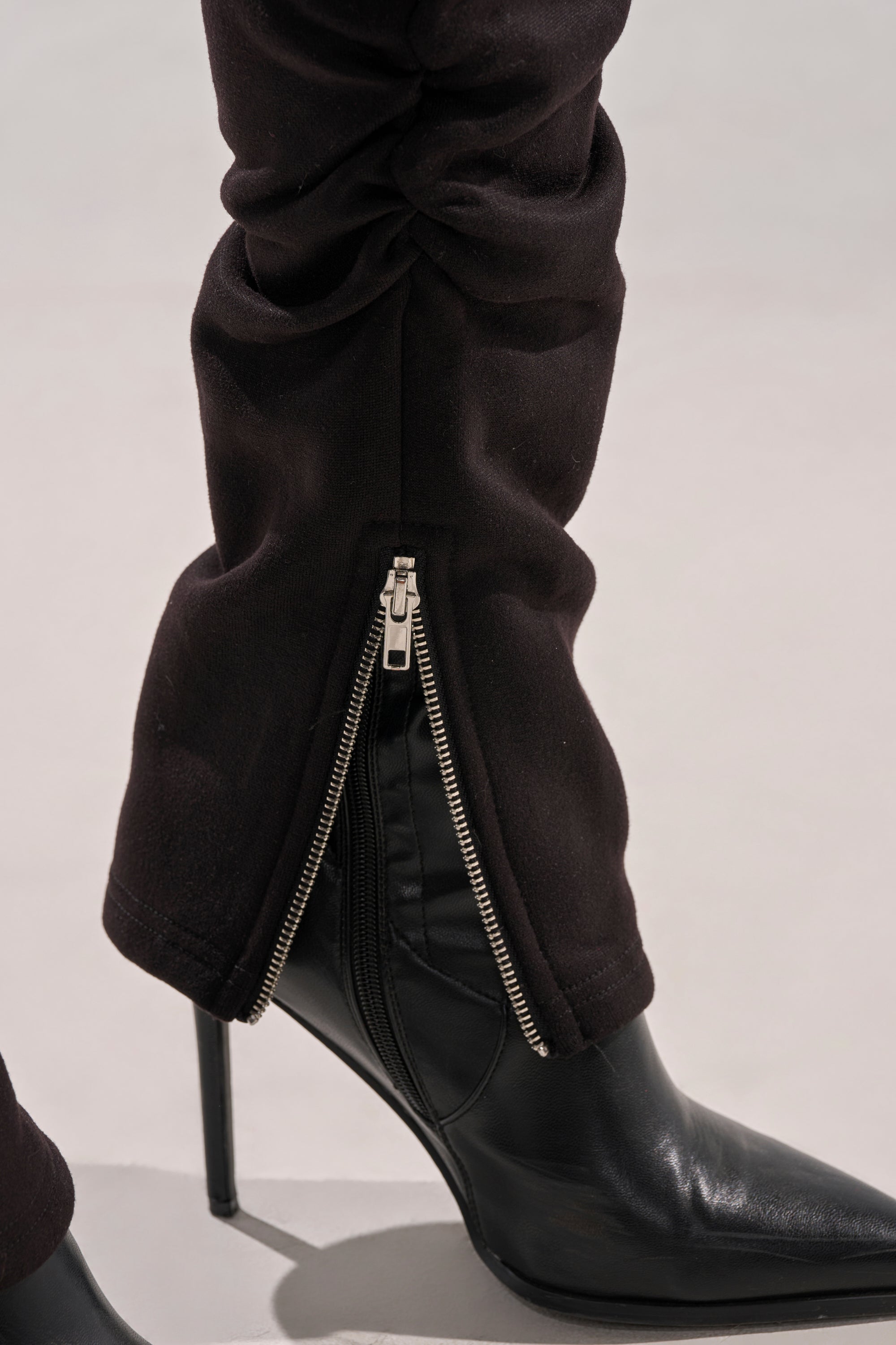 A close-up of the COZY HOLIDAY RHINESTONE JOGGER IN BLACK, with an open silver ankle zipper, partially covering a pointed black high-heeled boot. The background is a neutral light gray.