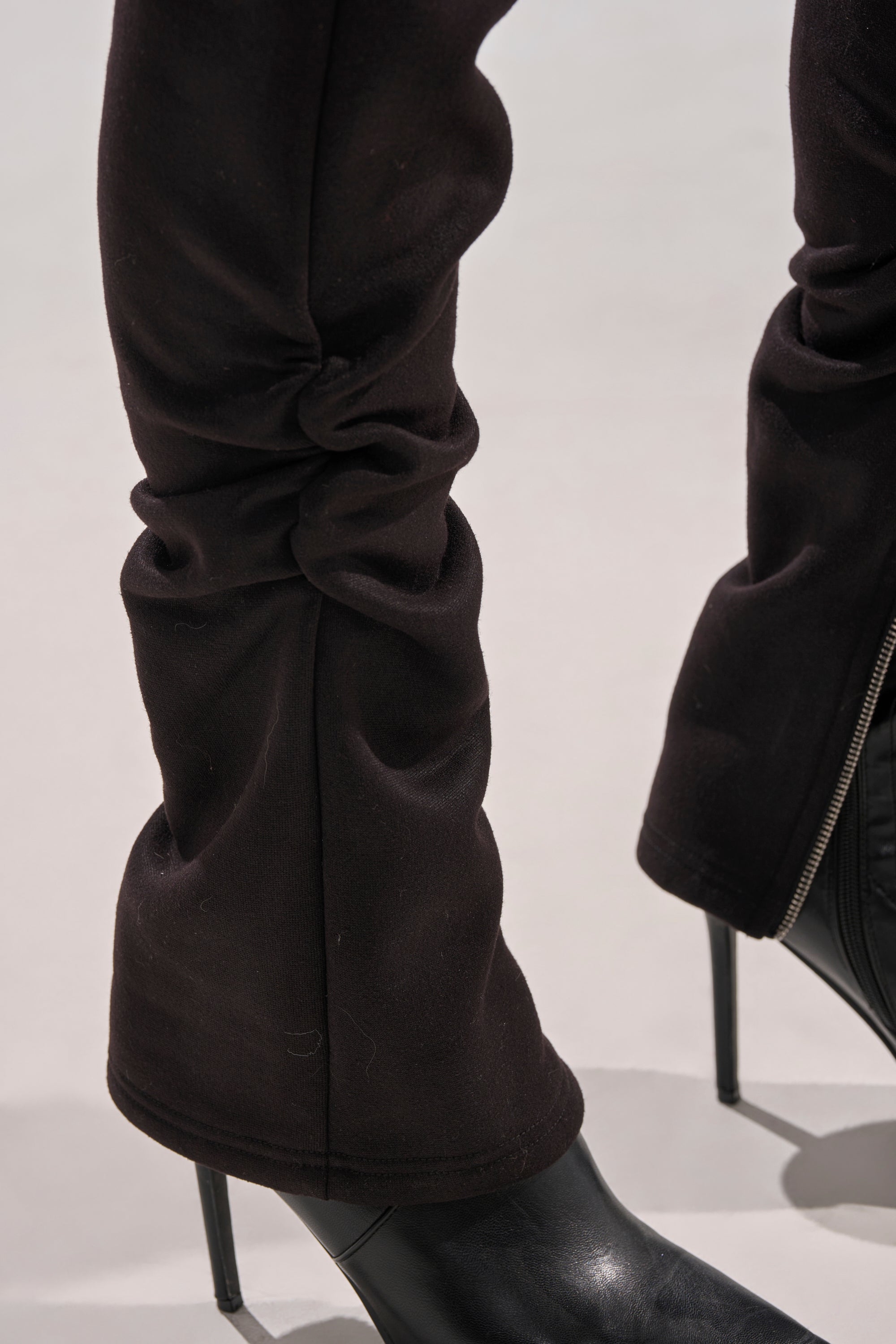 Close-up of a person wearing black heeled boots and the COZY HOLIDAY RHINESTONE JOGGER IN BLACK, featuring a zipper detail near the ankle, standing on a light-colored surface.