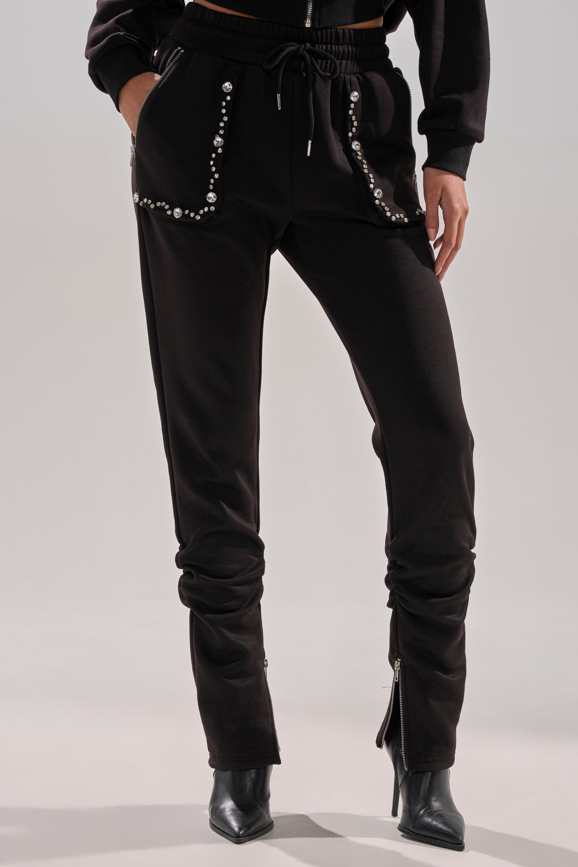 Wearing the COZY HOLIDAY RHINESTONE JOGGER IN BLACK with sparkling pocket accents, zipped ankles, and a matching sweatshirt, paired with black pointed-toe heeled boots for a chic holiday look.