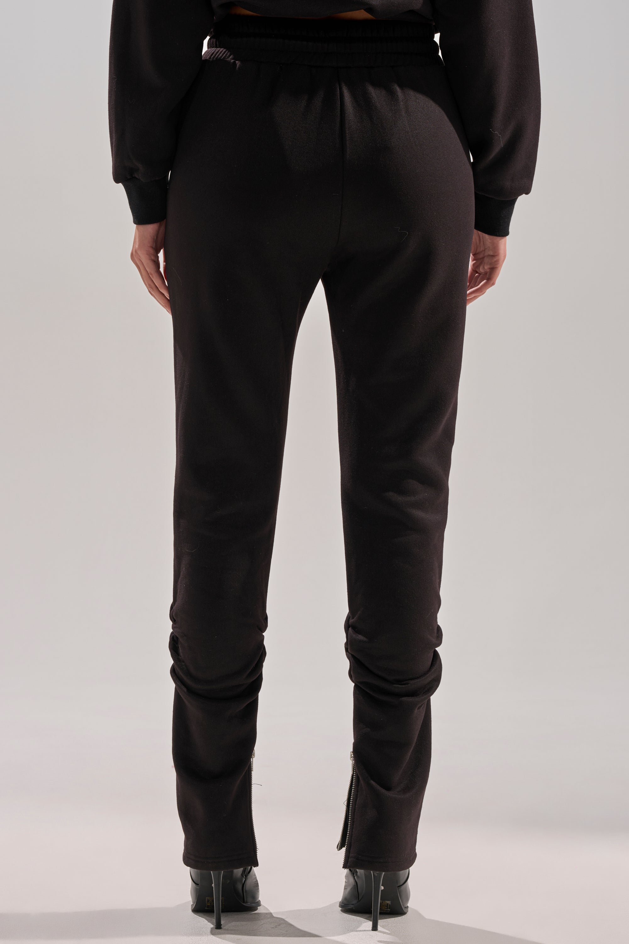 Person stands facing away from the camera, wearing the COZY HOLIDAY RHINESTONE JOGGER IN BLACK with a black long-sleeve top and black high-heeled shoes, showcasing visible zippers at the ankles.