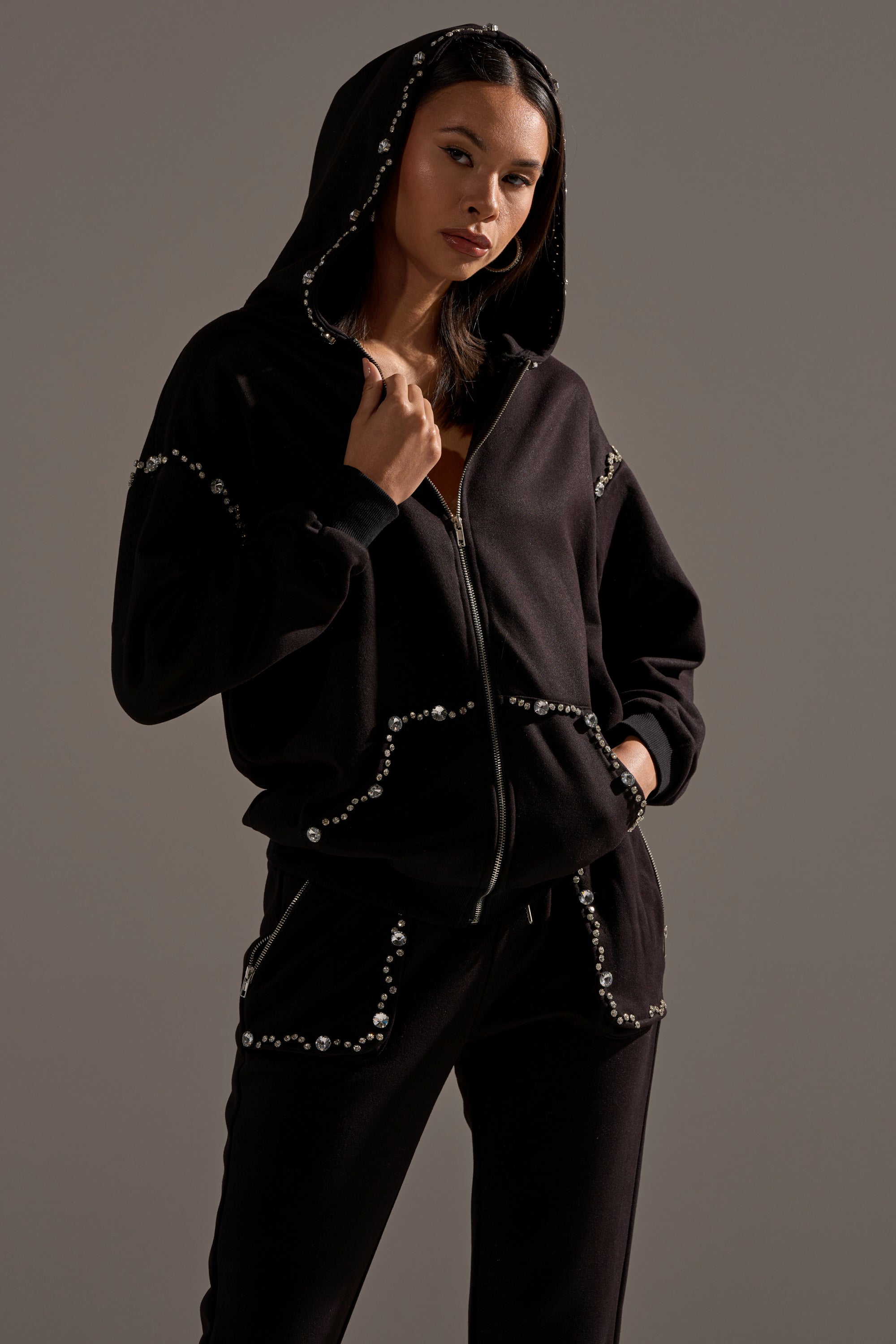A woman models the COZY HOLIDAY RHINESTONE ZIP UP SWEATSHIRT IN BLACK, paired with matching pants featuring rhinestone details, as she poses confidently with one hand on her hood against a plain background.