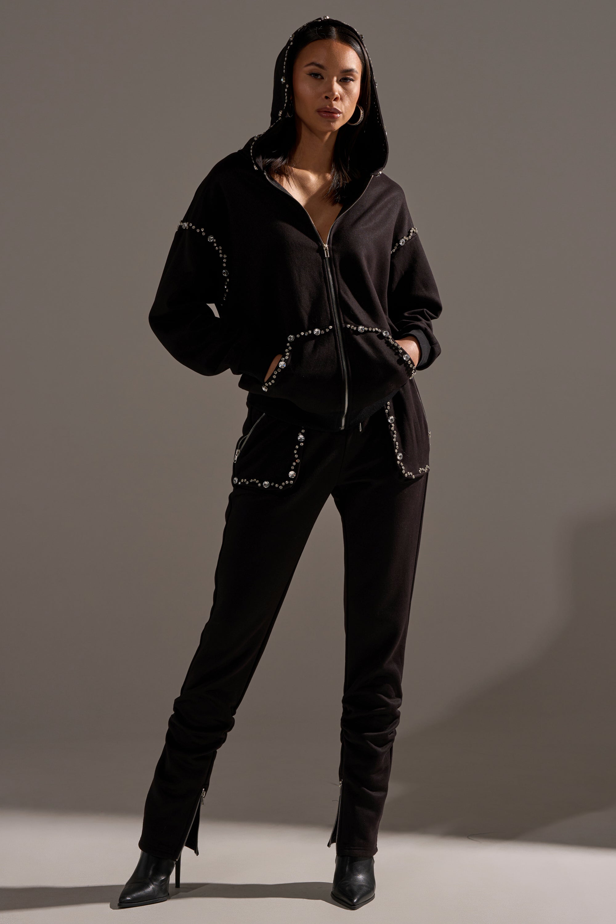 A woman stands confidently in the AKIRA Label COZY HOLIDAY RHINESTONE ZIP UP SWEATSHIRT IN BLACK and matching pants, featuring silver embellishments, paired with black heeled boots against a neutral gray background.