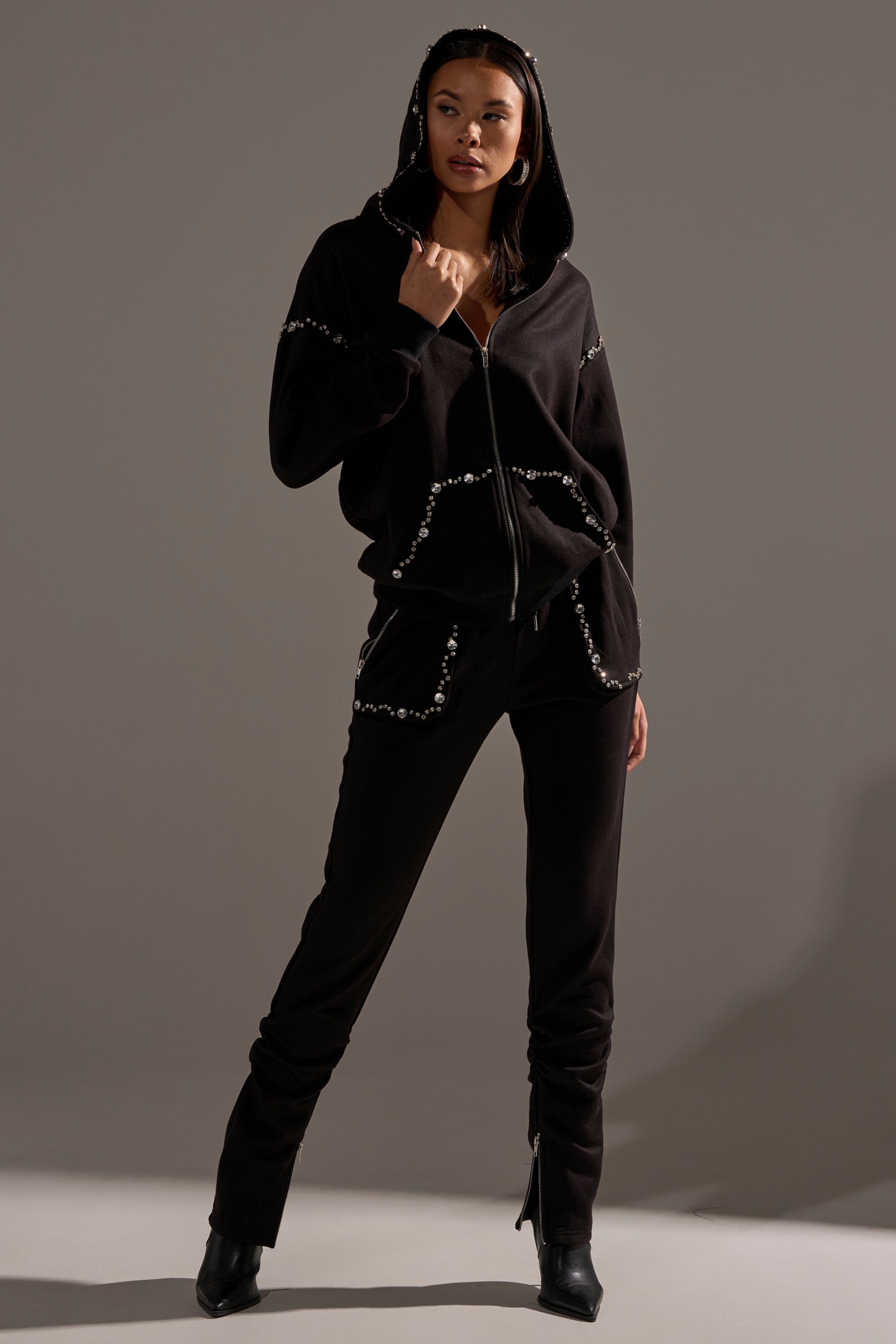 A woman stands confidently with one hand in her pocket, wearing the COZY HOLIDAY RHINESTONE ZIP UP SWEATSHIRT IN BLACK and black pants with pearl-like beads, against a plain gray background.