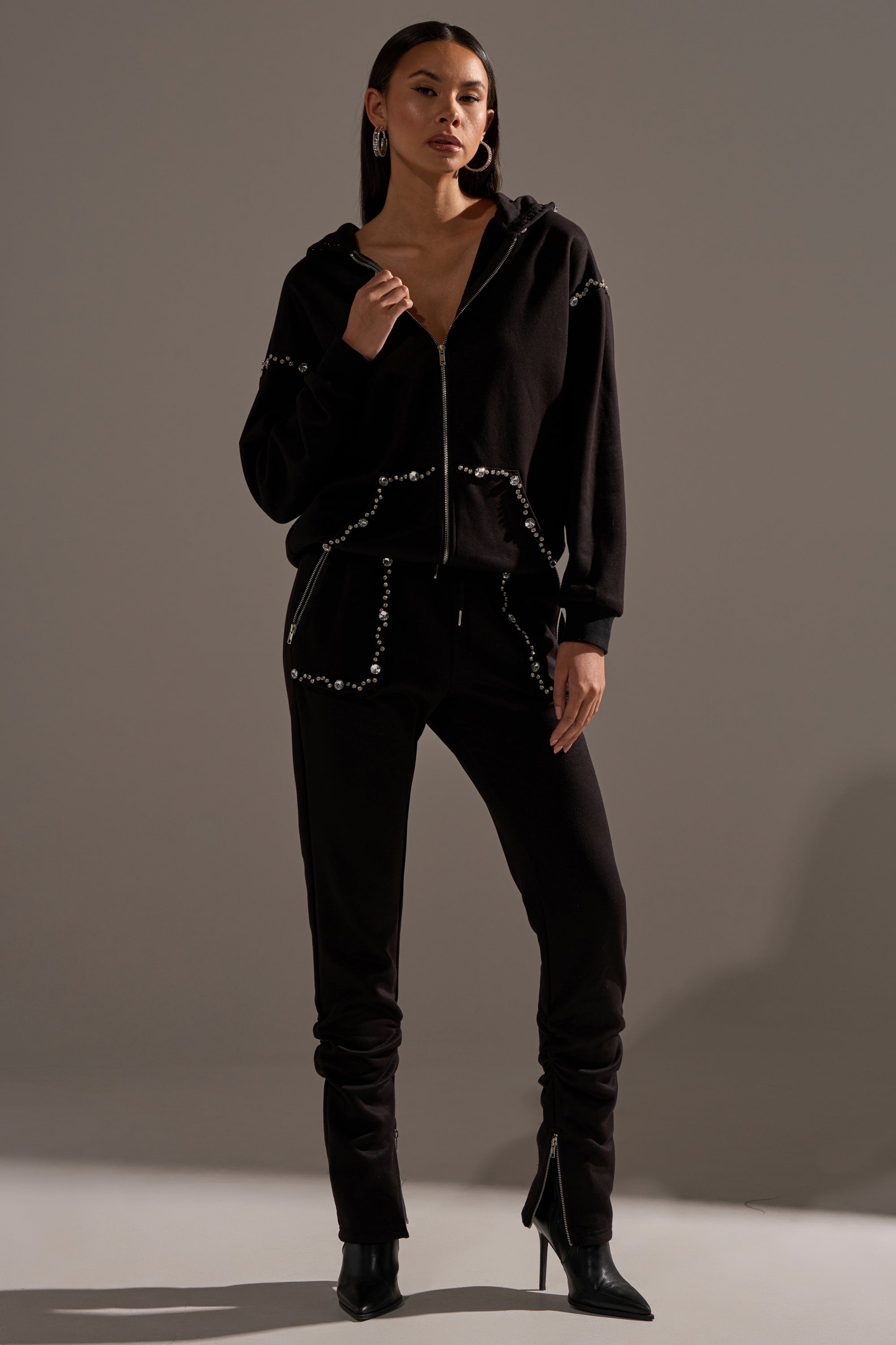 A woman stands confidently in the COZY HOLIDAY RHINESTONE ZIP UP SWEATSHIRT IN BLACK, paired with matching pants and black heeled boots, posing against a plain gray background.