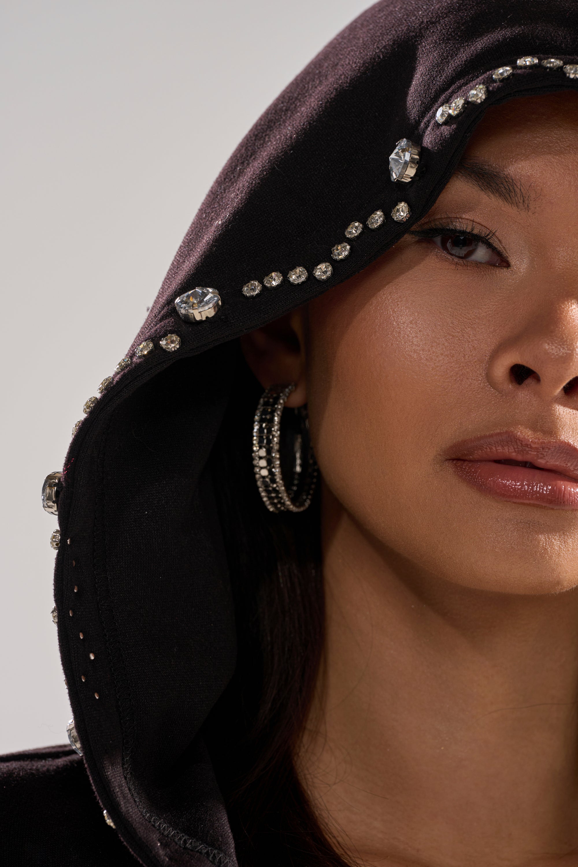 Close-up of a woman wearing the COZY HOLIDAY RHINESTONE ZIP UP SWEATSHIRT IN BLACK. Only the right side of her face is visible, showcasing the sparkling rhinestone details on the chic oversized hoodie. She has glossy lips, hoop earrings, and smooth skin.